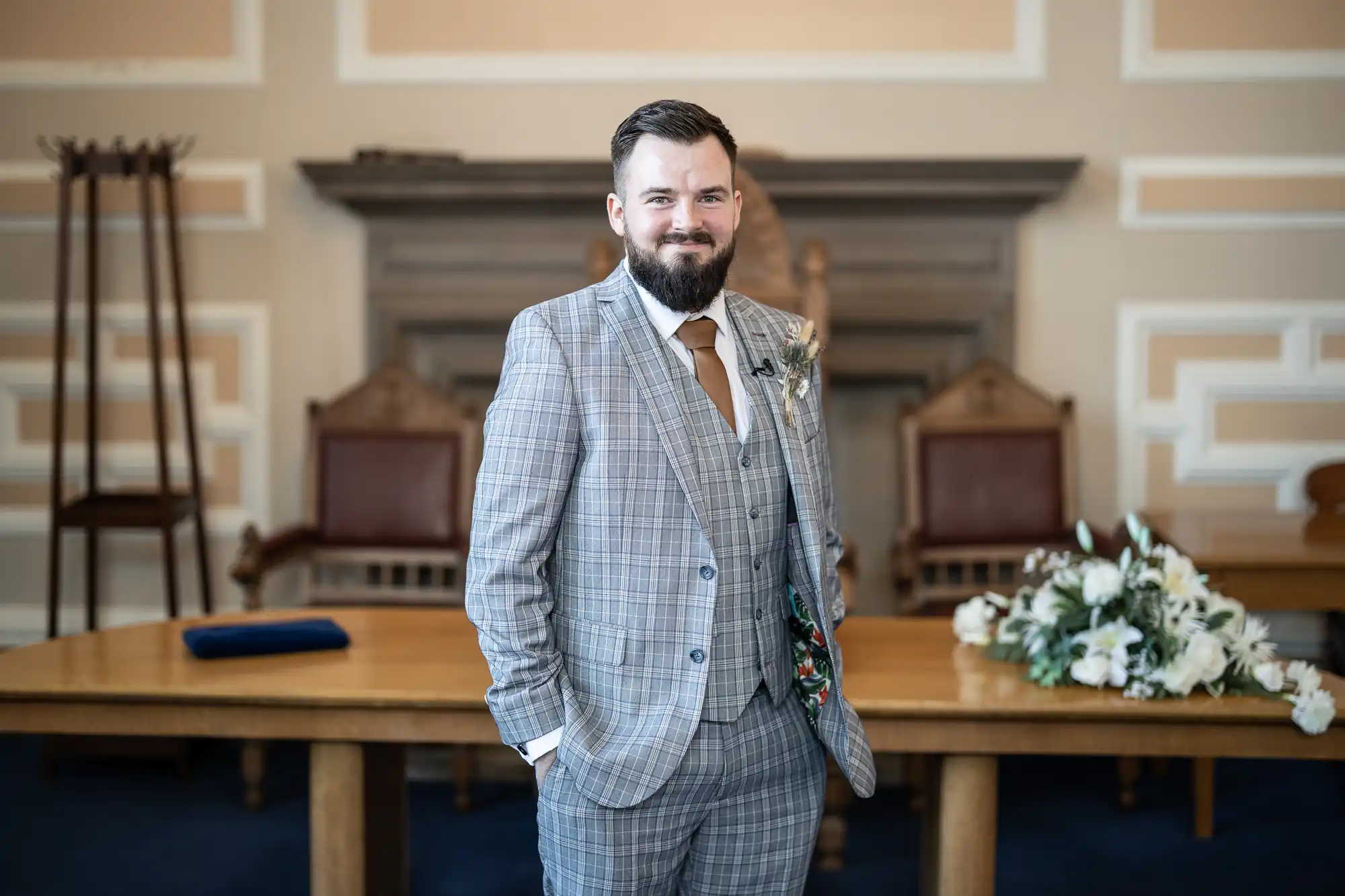 A man with a beard, wearing a grey checked suit, tan tie, and buttonhole, stands in a formal room with wooden furniture, floral arrangement, and intricate wall moulding. He smiles slightly, hands in pockets, next to a large wooden table.