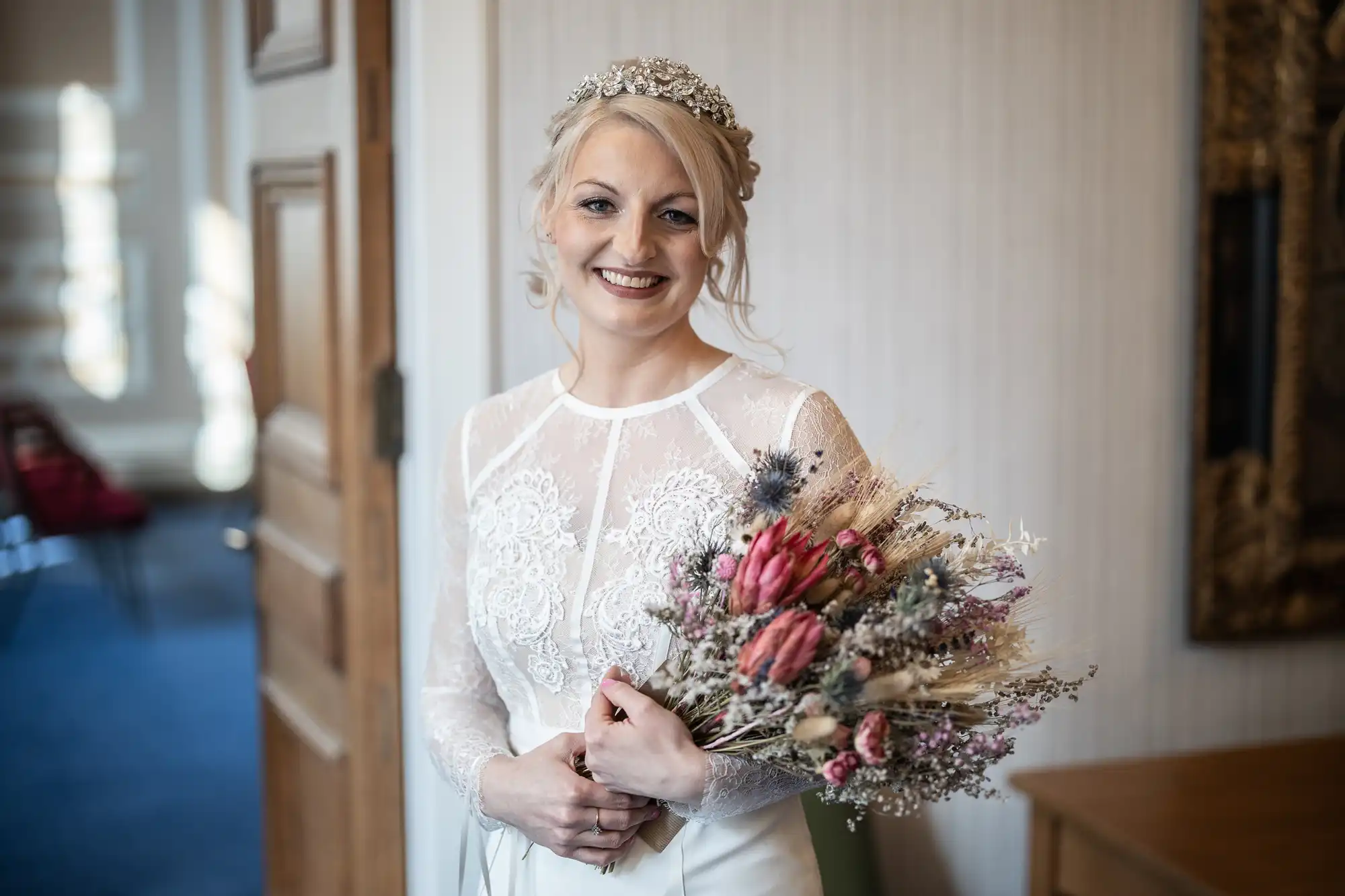 A smiling bride with blonde hair and a jewelled headpiece stands indoors, wearing a long-sleeved lace wedding dress and holding a rustic bouquet of dried flowers in pink, purple, and cream tones. A wooden door and ornate gold frame are visible in the background.