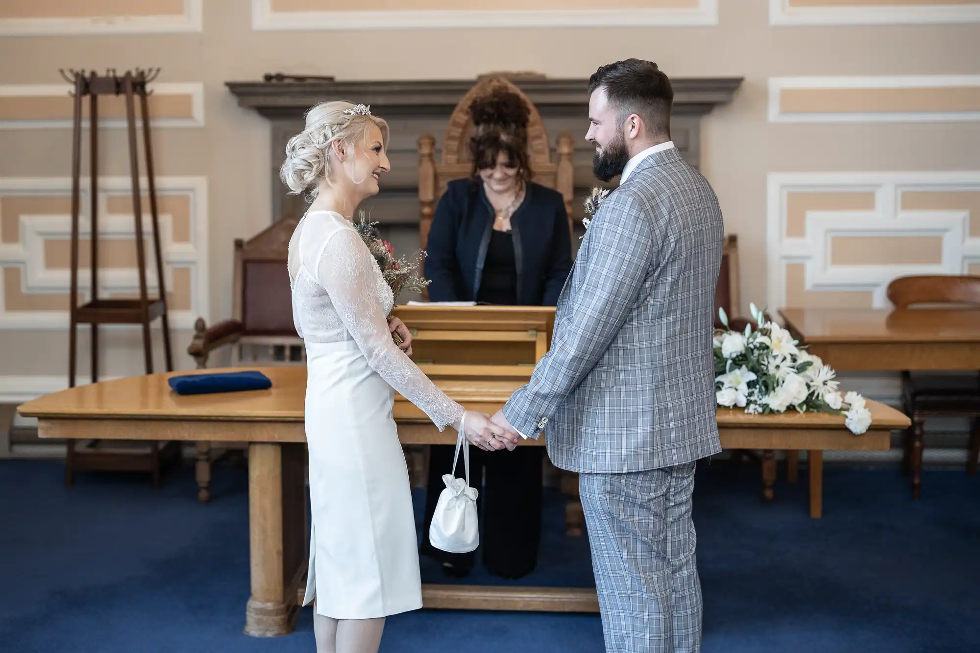 A bride and groom stand holding hands, facing each other and smiling, in a formal room with blue carpet. The bride wears a white dress and holds a small white bag; the groom wears a grey checked suit. An officiant stands behind them at a wooden table with flowers.
