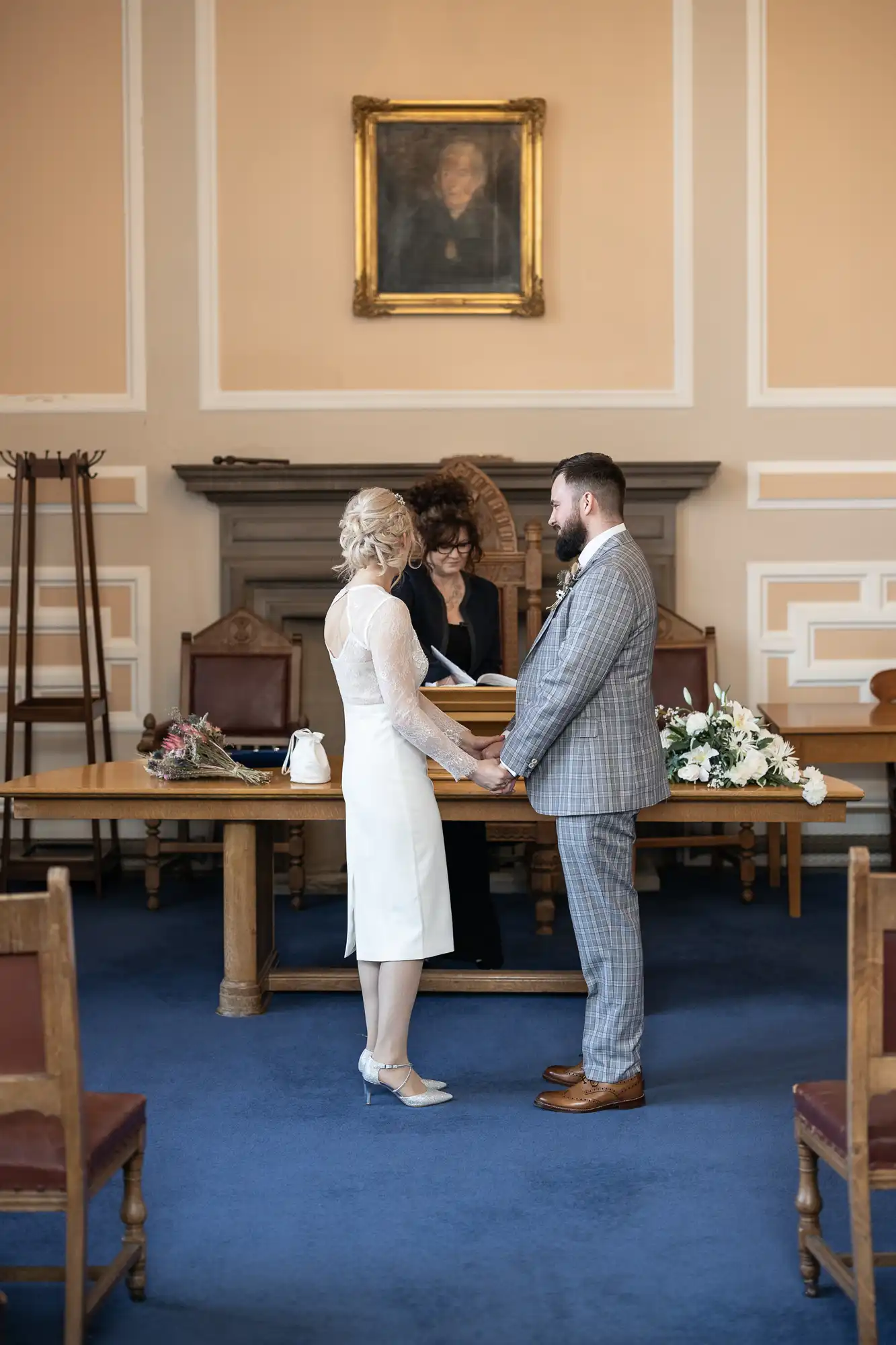 A bride and groom stand facing each other, holding hands in a formal room with blue carpet and ornate cream walls. An officiant stands behind a wooden table with flowers and books. Empty chairs line the sides, and a portrait hangs above the scene.