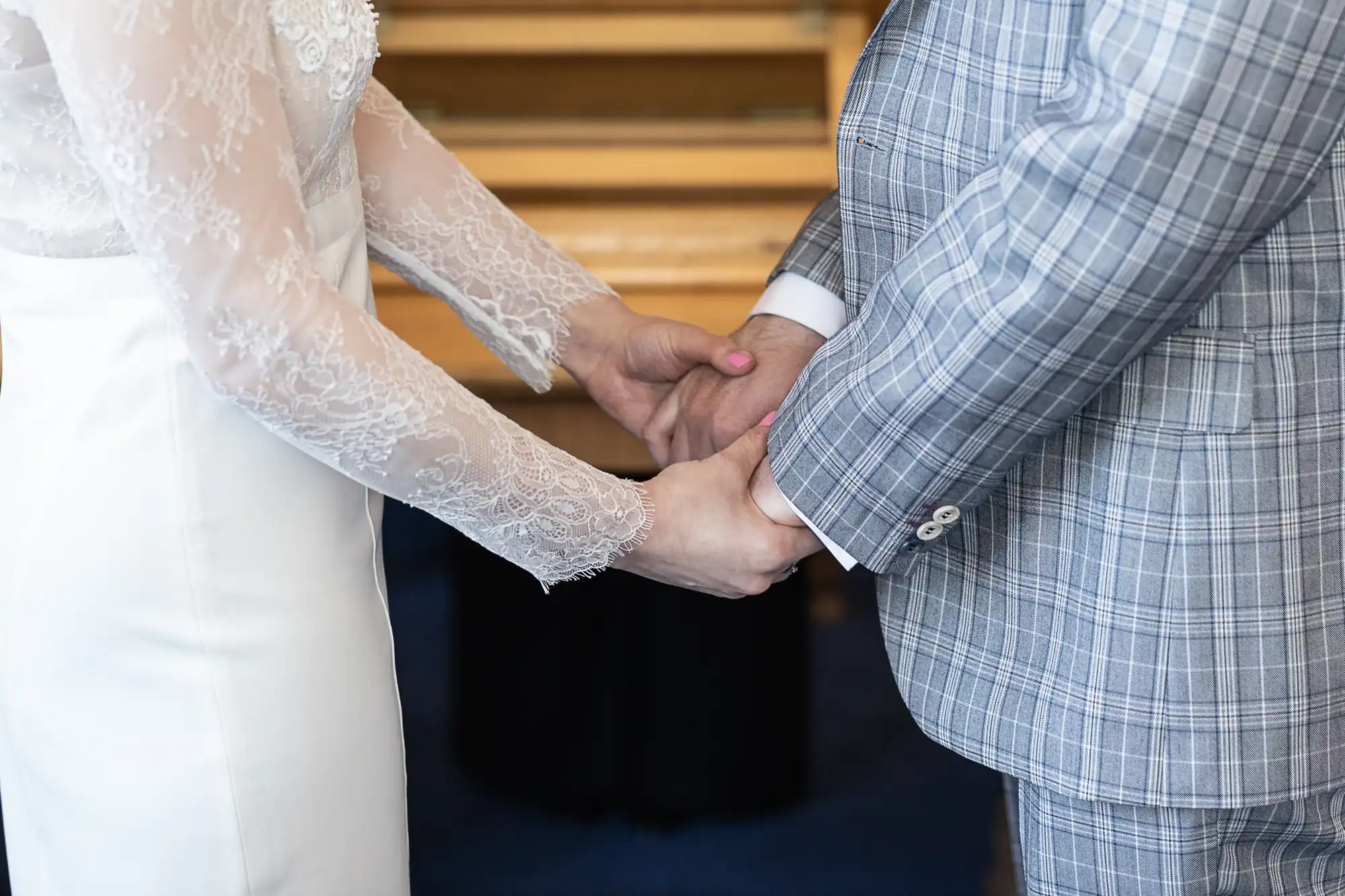 A couple hold hands indoors. One person wears a white dress with lace sleeves; the other wears a light grey checked suit. The scene suggests a formal or wedding ceremony. Only their bodies from shoulders to waist are visible, not their faces.