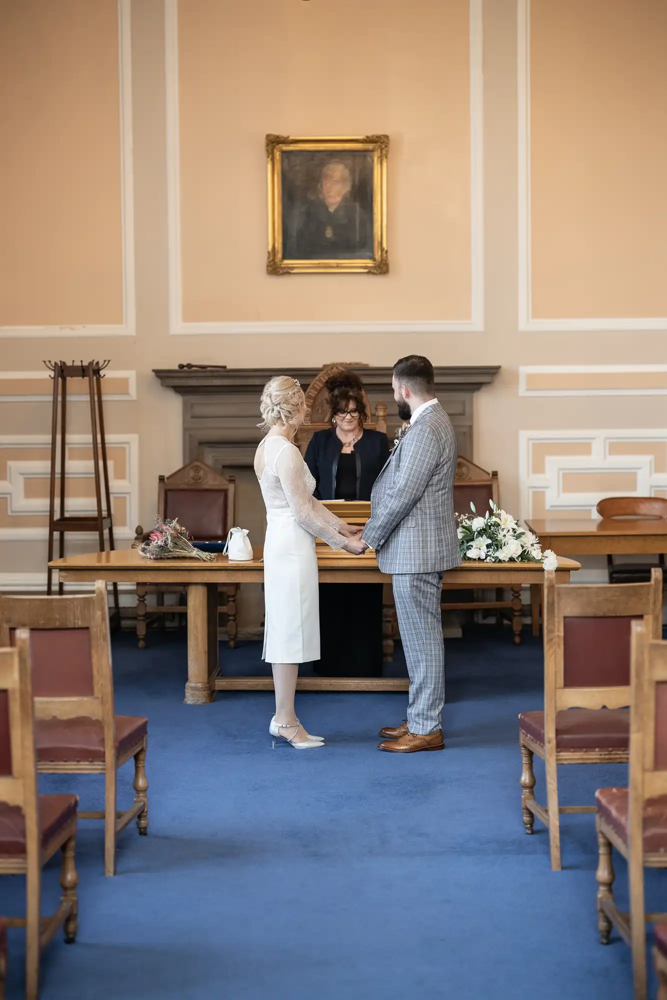A bride and groom stand facing each other, holding hands in a small, elegant room with blue carpet. A celebrant stands behind a wooden table adorned with flowers. Empty chairs line the room, and a portrait hangs on the beige wall behind them.