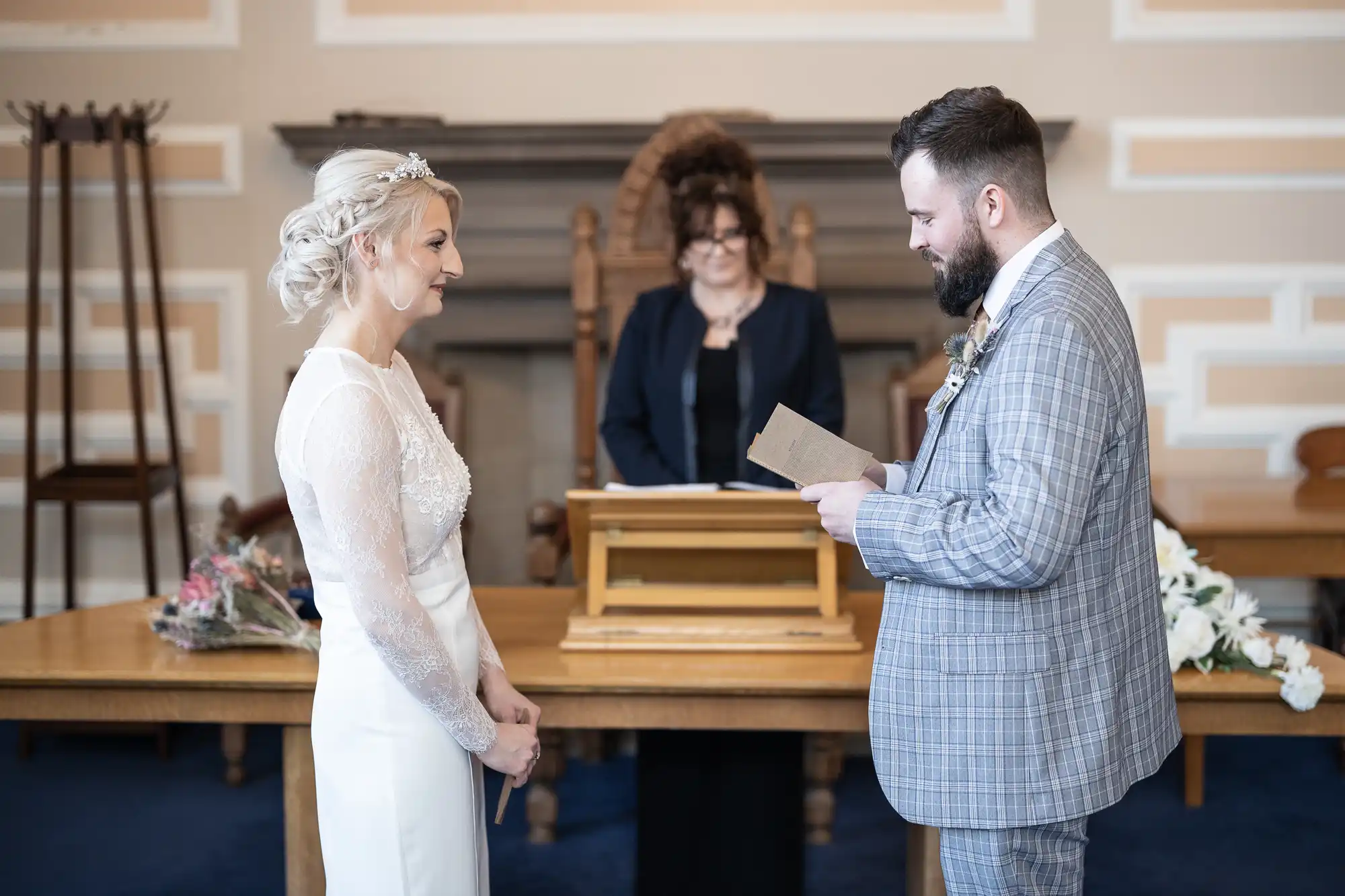 A bride in a white dress and a groom in a grey checked suit stand facing each other, smiling, as the groom reads from a card. An officiant stands behind a wooden lectern in a formal room with pale walls and blue carpet. A bouquet rests on the table.