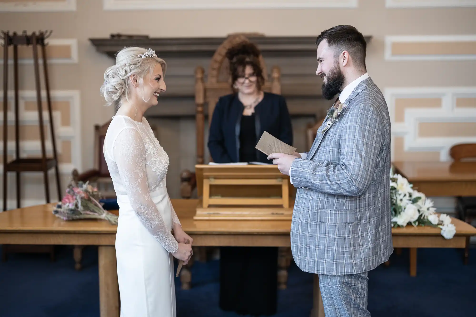 A bride and groom face each other, smiling, during a wedding ceremony. The groom, holding a piece of paper, wears a grey checked suit, while the bride wears a white dress with lace sleeves. An officiant stands behind a desk, flowers and papers visible on the table.