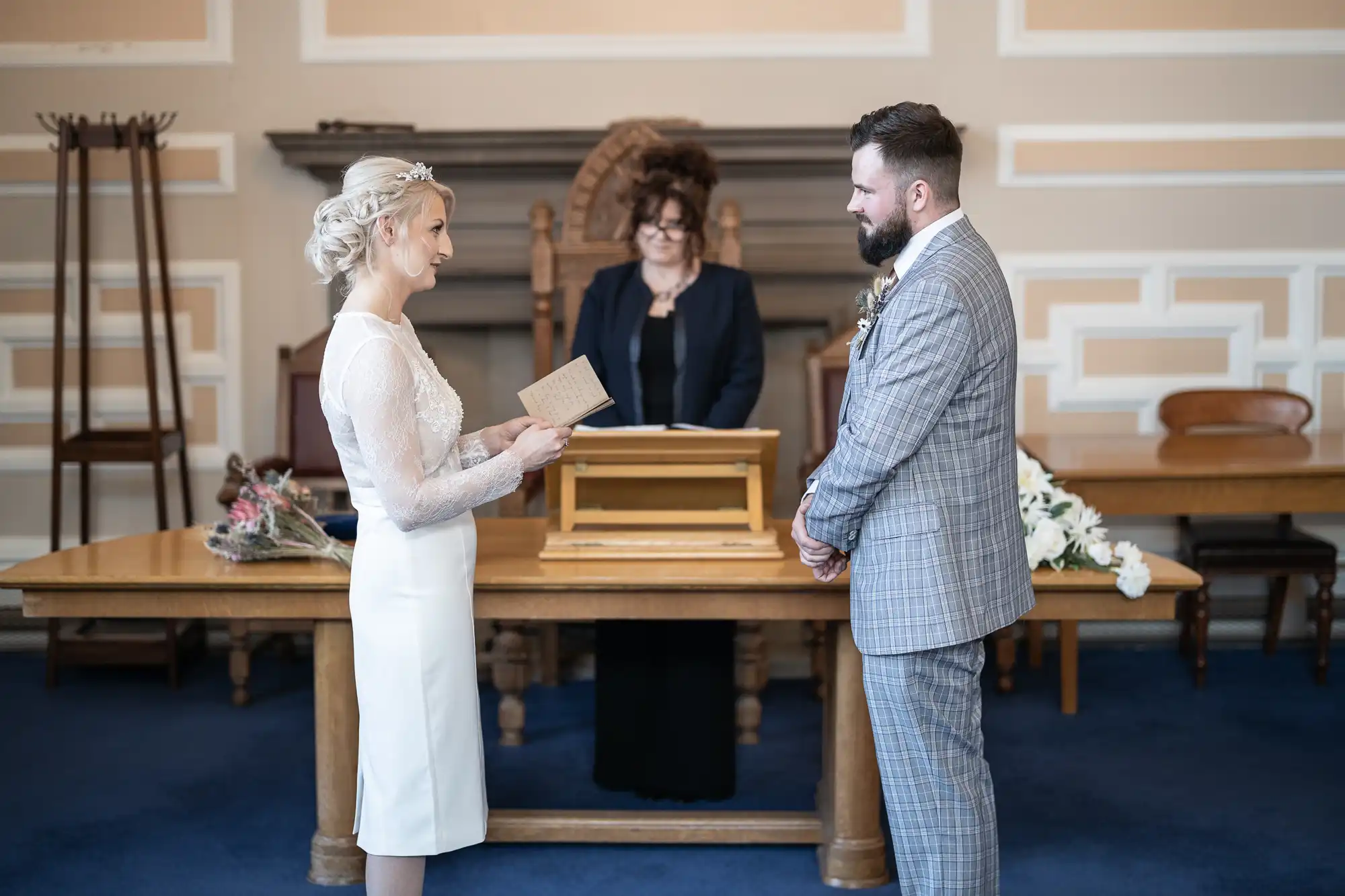A bride in a white dress reads from a card to a groom in a checked suit. They face each other, standing in front of a table, with a bouquet nearby. An officiant stands behind the table in a formal setting with wooden furniture and decorative walls.