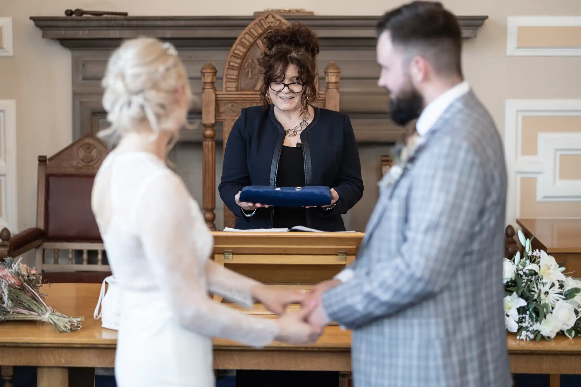 A bride and groom hold hands facing each other in the foreground, both slightly out of focus. In the background, a smiling celebrant stands behind a wooden lectern, holding a blue folder. The setting appears formal, with floral arrangements on the table.