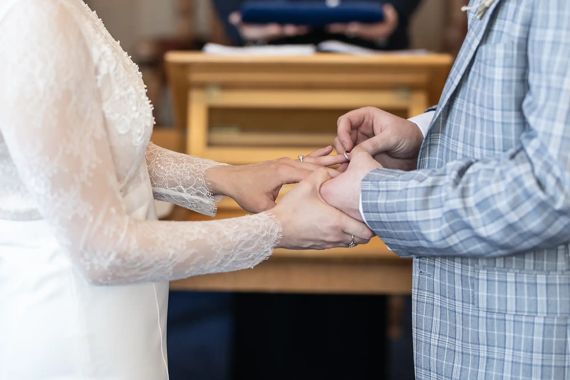 A close-up of two people exchanging rings during a wedding ceremony. One person wears a white lace dress, and the other wears a light checked suit. Their hands are gently touching, and a wooden table is blurred in the background.