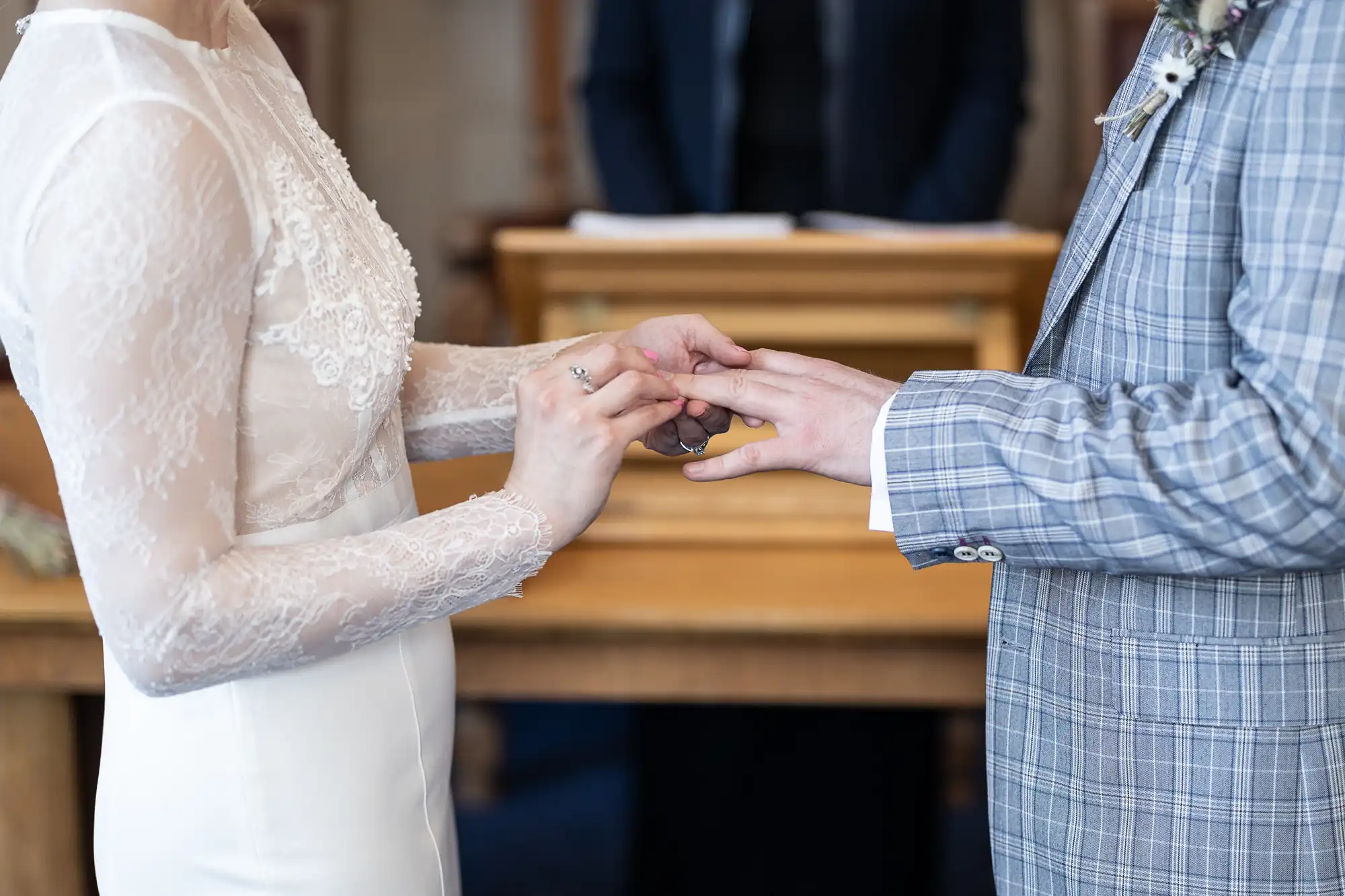A bride in a white lace dress places a ring on the groom’s finger. The groom wears a light checked suit. They stand close together indoors, with a wooden table and a blurred figure in the background. Only their torsos and hands are visible.