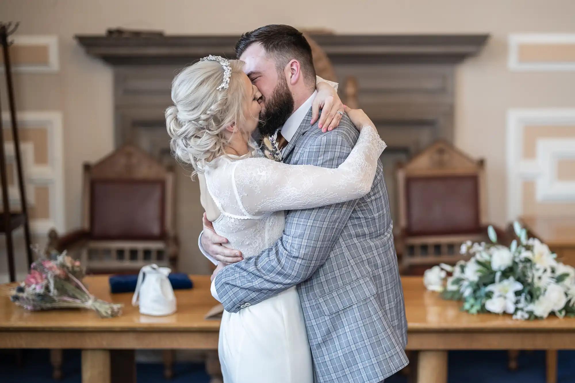 A bride and groom embrace and kiss indoors, both dressed formally. The bride wears a white dress and jewelled headpiece; the groom wears a checked grey suit. They stand before a wooden table with flowers, with ornate chairs and a fireplace in the background.
