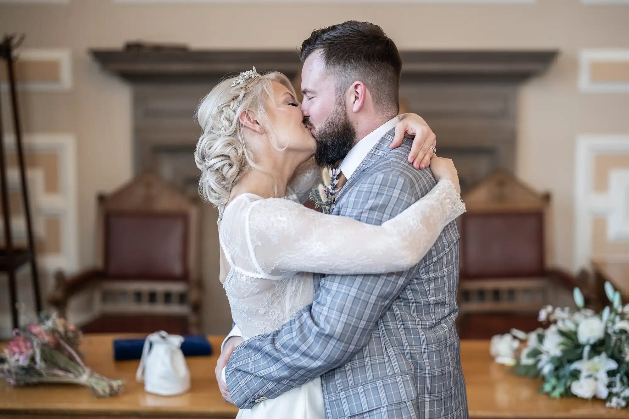 A bride and groom share a kiss at their wedding. The bride wears a lace-sleeved white dress and the groom wears a grey checked suit. They embrace in front of a wooden table with flowers and vintage chairs in the background.