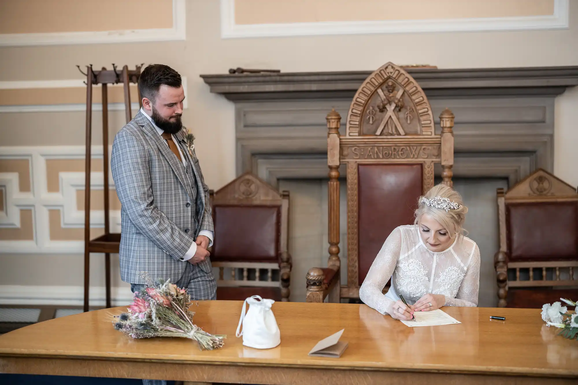 A bride in a white lace dress signs a document at a wooden table, while a groom in a grey checked suit stands nearby, watching. A bouquet and handbag rest on the table. Ornate chairs and a marble fireplace are in the background.