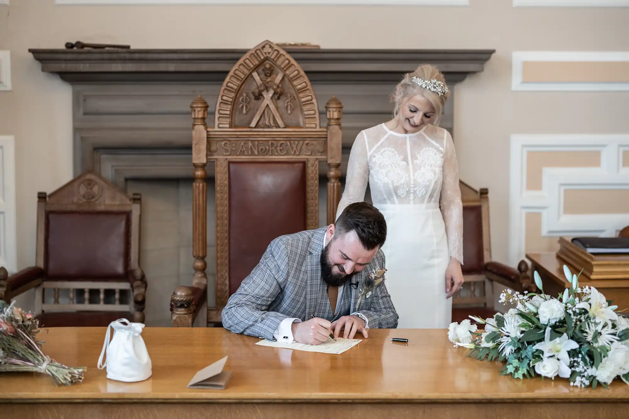 A man in a chequered suit sits at a wooden desk, signing a document whilst smiling. Standing behind him, a woman in a long-sleeved, white lace wedding dress smiles down at him. They are in a formal room with large chairs, flowers, and ornate woodwork.