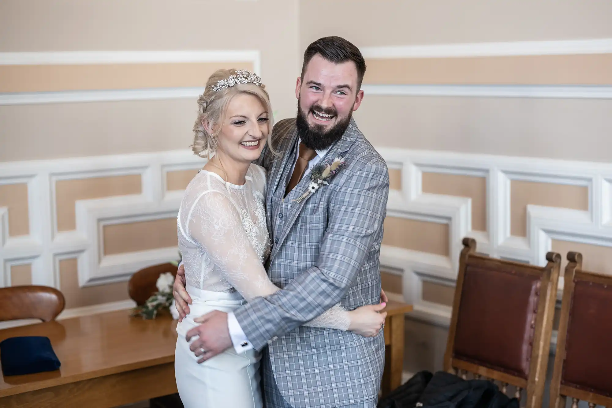 A bride in a white lace dress and tiara embraces a groom in a grey checked suit with a buttonhole. Both are smiling joyfully. They stand in a room with cream walls, wooden chairs, and a table in the background.