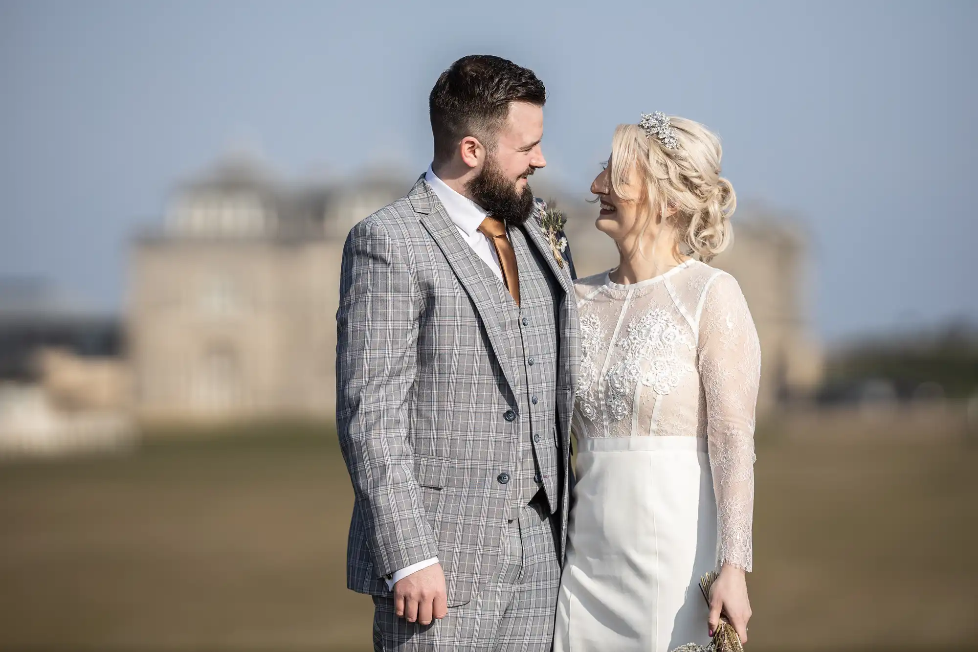 A groom in a grey checked suit and a bride in a white lace dress smile and gaze at each other outdoors, with an elegant, blurred building in the background on a sunny day.