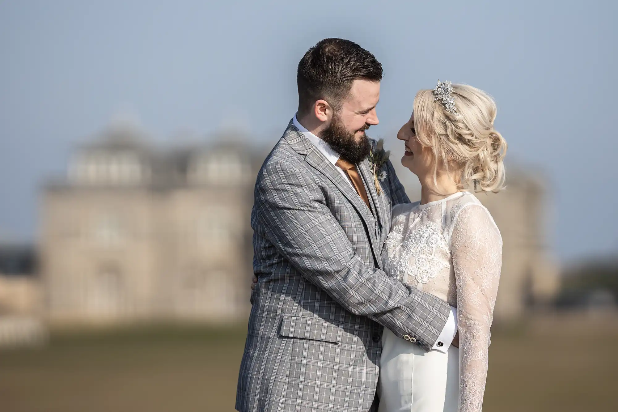 A groom in a grey checked suit embraces a bride in a long-sleeved lace wedding dress. They are outdoors, gazing and smiling at each other, with a large, blurred historic building in the background under a clear sky.
