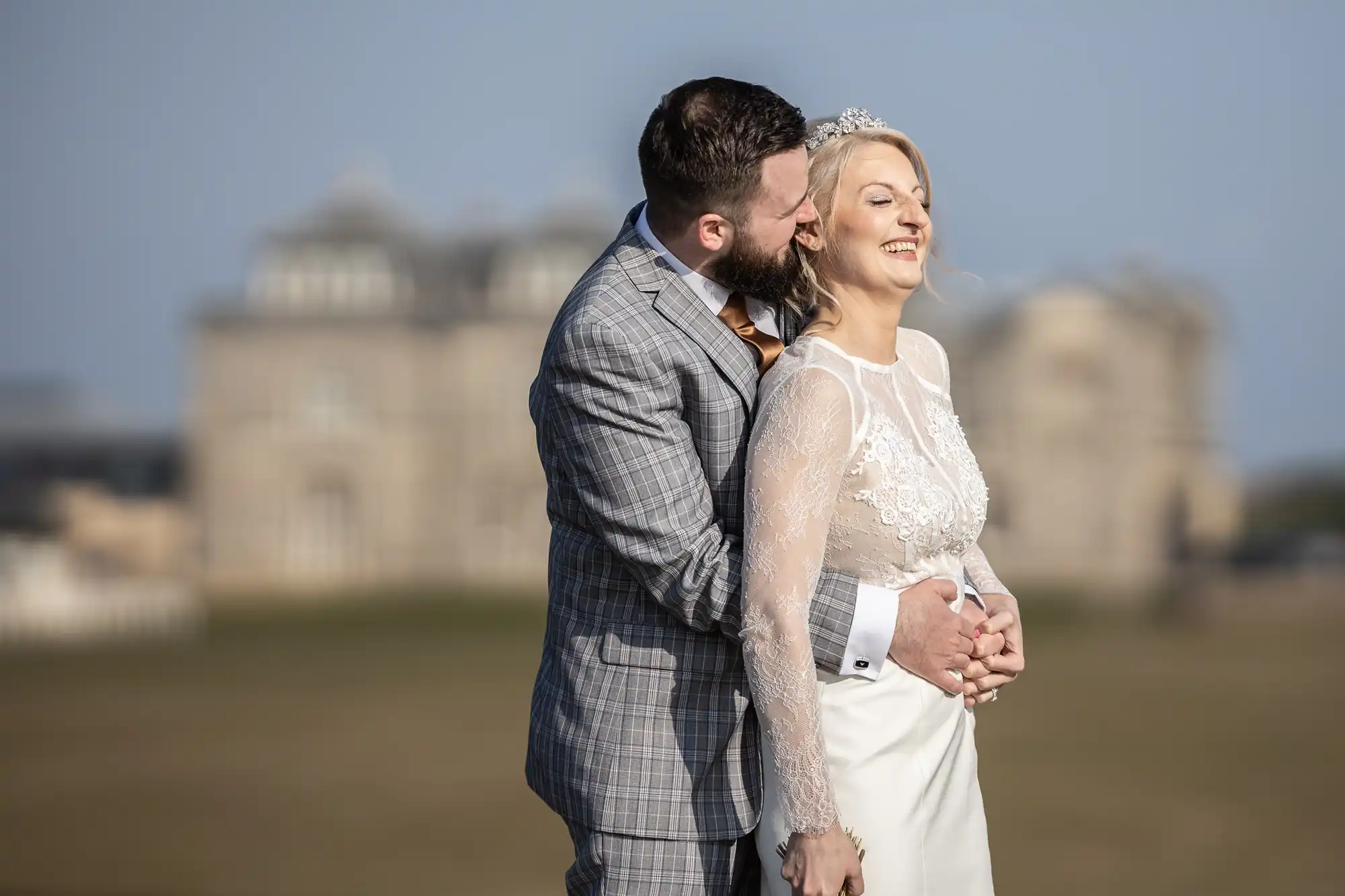 A smiling bride in a white lace dress stands in front of a groom in a grey checked suit who embraces her from behind and kisses her cheek. They are outdoors with a blurred historic building in the background under a clear sky.
