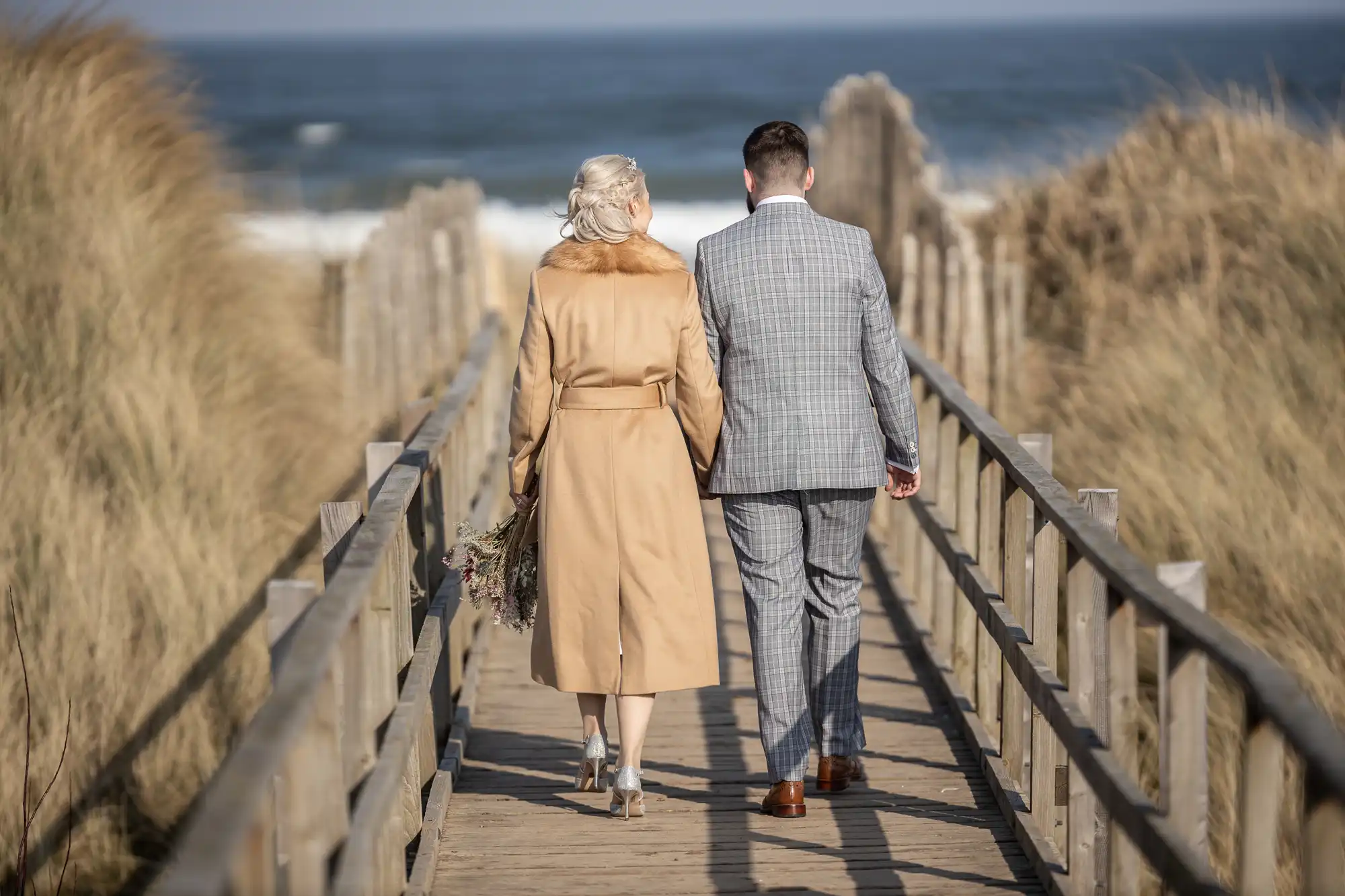 A couple walks hand-in-hand on a wooden boardwalk towards the beach. The woman in a tan coat holds a bouquet, and the man wears a grey checked suit. Tall grasses border the boardwalk, and the sea is visible under a clear blue sky.