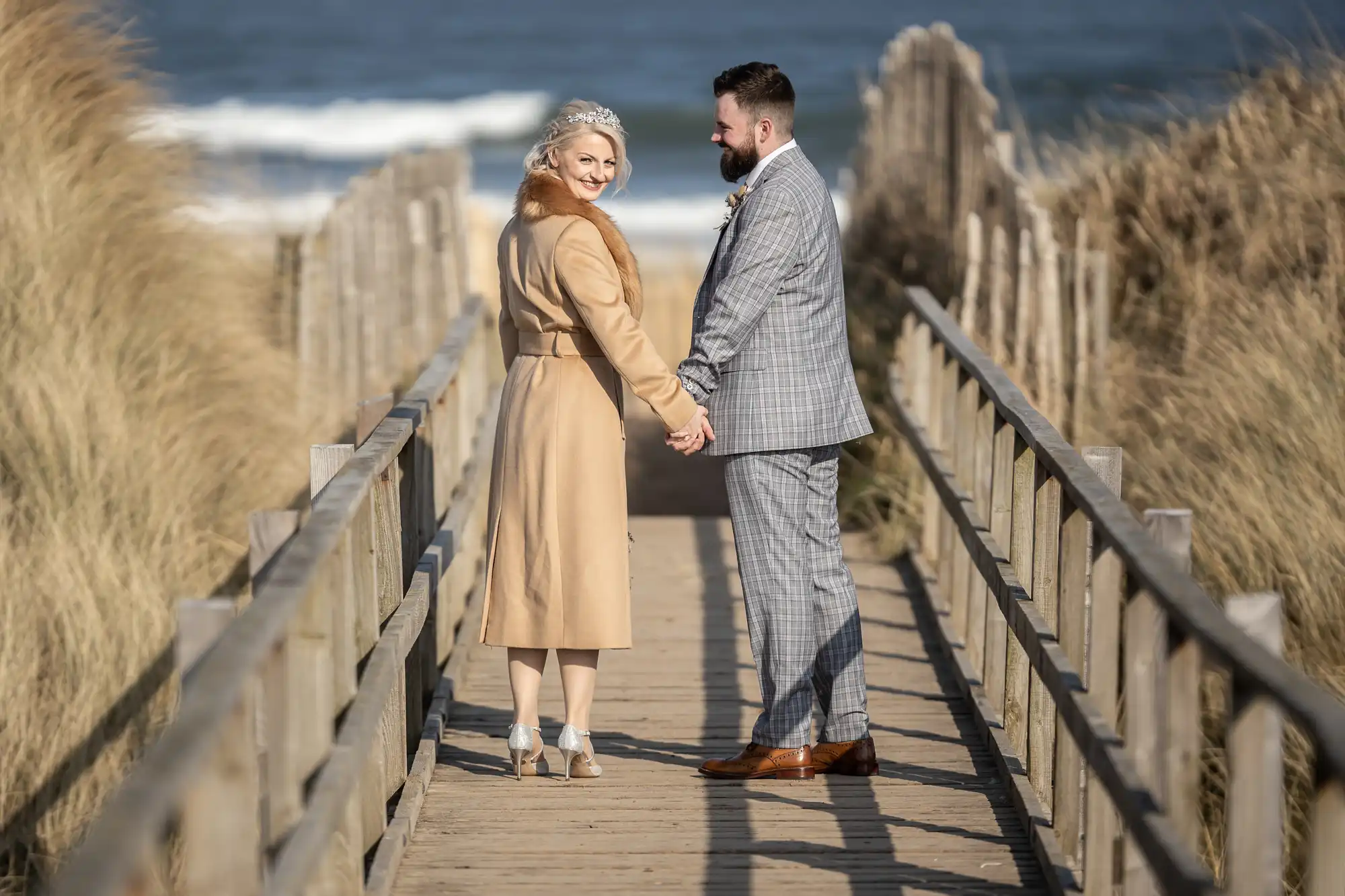 A couple stands on a wooden boardwalk leading to a beach, holding hands and smiling at the camera. The woman wears a beige coat and heels; the man wears a grey checked suit with brown shoes. Tall grass lines the sides; waves and blue sky are visible in the background.