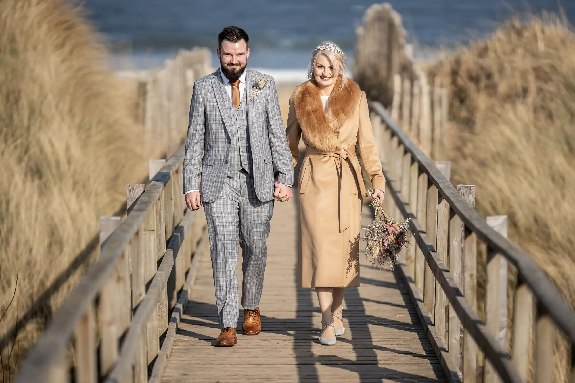 A couple walks hand-in-hand on a wooden boardwalk by the beach. The man wears a grey checked suit and brown shoes; the woman wears a tan coat with a fur collar and a jewelled headband, holding a bouquet. Grass and sand dunes line the path, with the sea in the background.