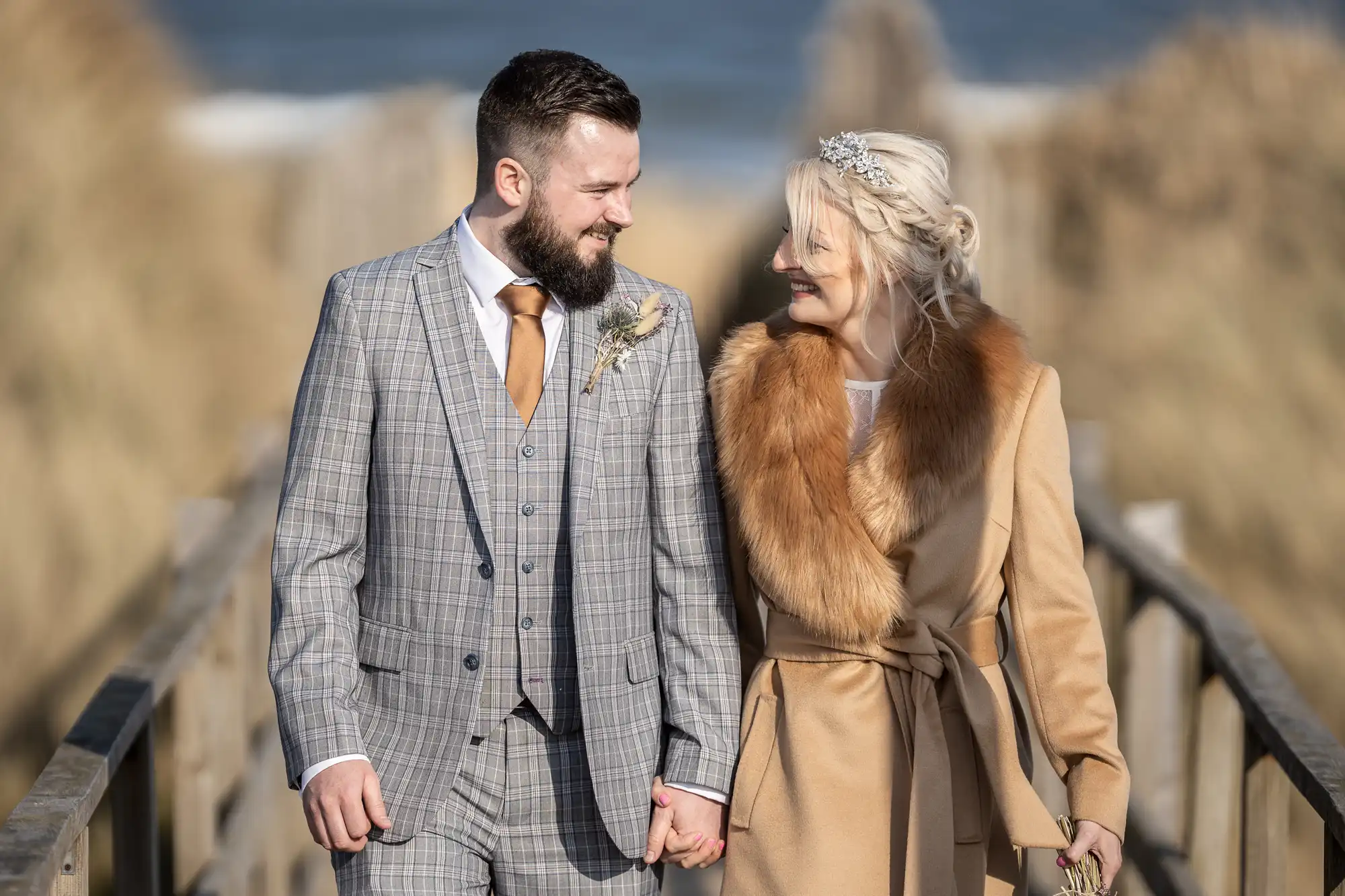 A couple walks outdoors on a wooden path. The man, with a beard, wears a grey checked suit and tie, holding hands with the woman, who has fair hair and wears a camel coat with a fur collar and a decorative headband. Both are smiling at each other.