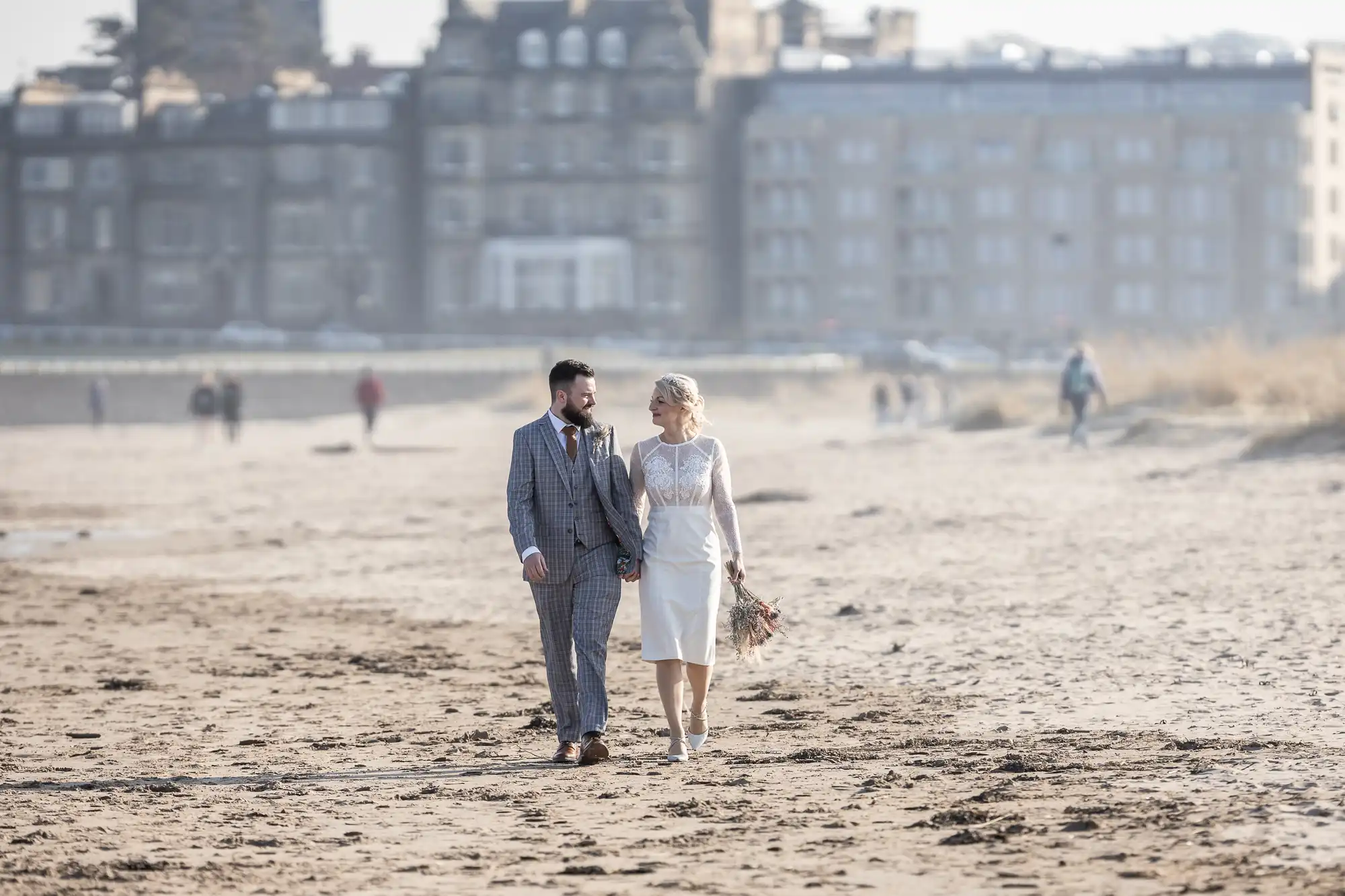 A couple in wedding attire walk hand in hand along a sandy beach. The groom wears a checked suit, and the bride wears a knee-length white dress, holding a bouquet. The background shows a large, blurred building and a few distant people.