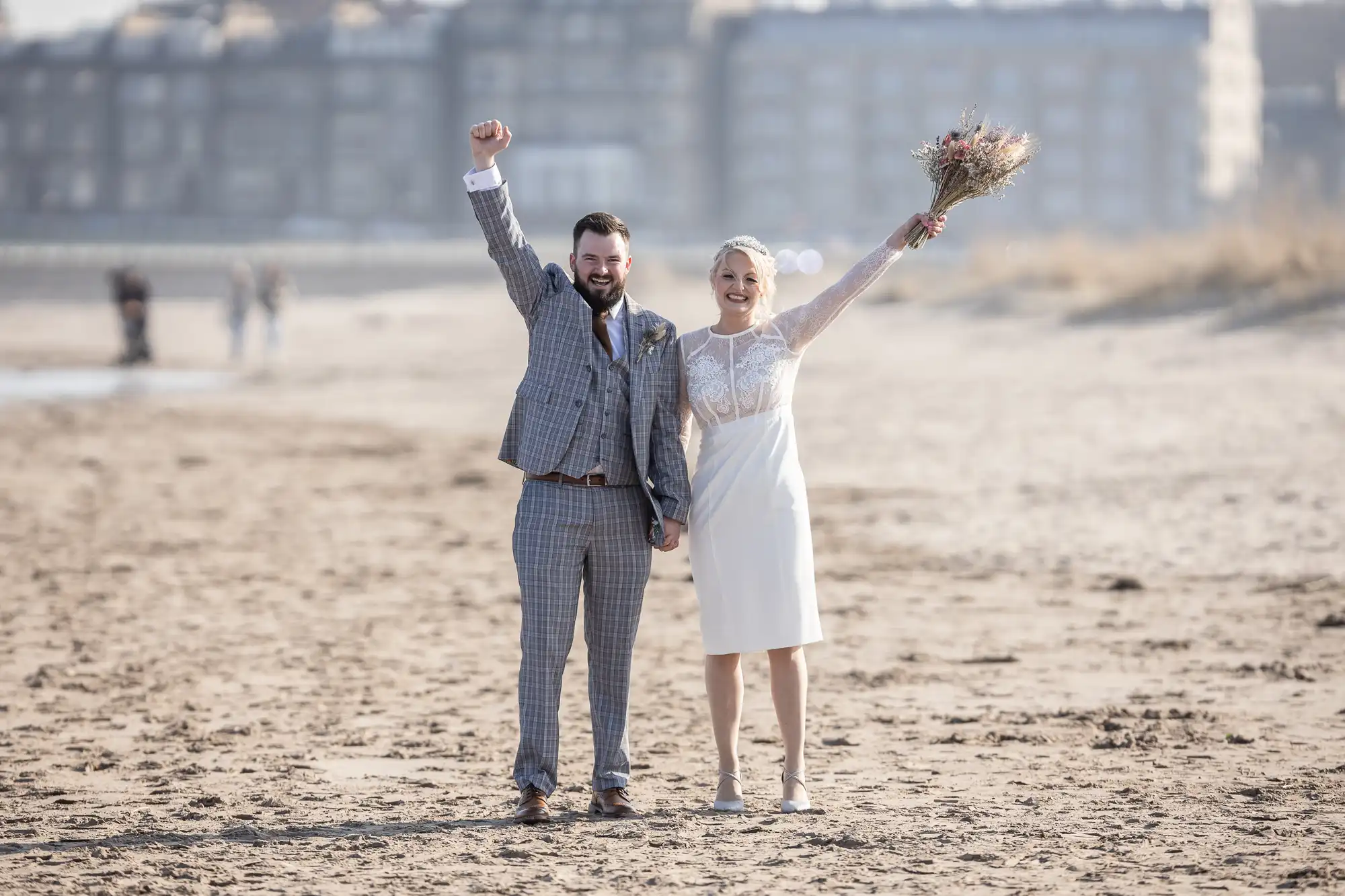 A joyful couple stands on a sandy beach, holding hands. The man in a grey checked suit raises his fist triumphantly. The woman in a white dress and lace top holds a bouquet aloft, both smiling. Blurred buildings and people are visible in the background.