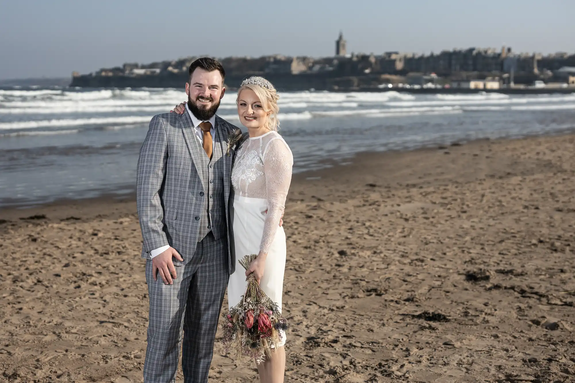 A smiling bride and groom stand arm in arm on a sandy beach. The groom wears a checked grey suit, brown tie, and white shirt. The bride wears a white dress with lace sleeves and a headpiece, holding a bouquet. Waves and a distant town are visible behind them.