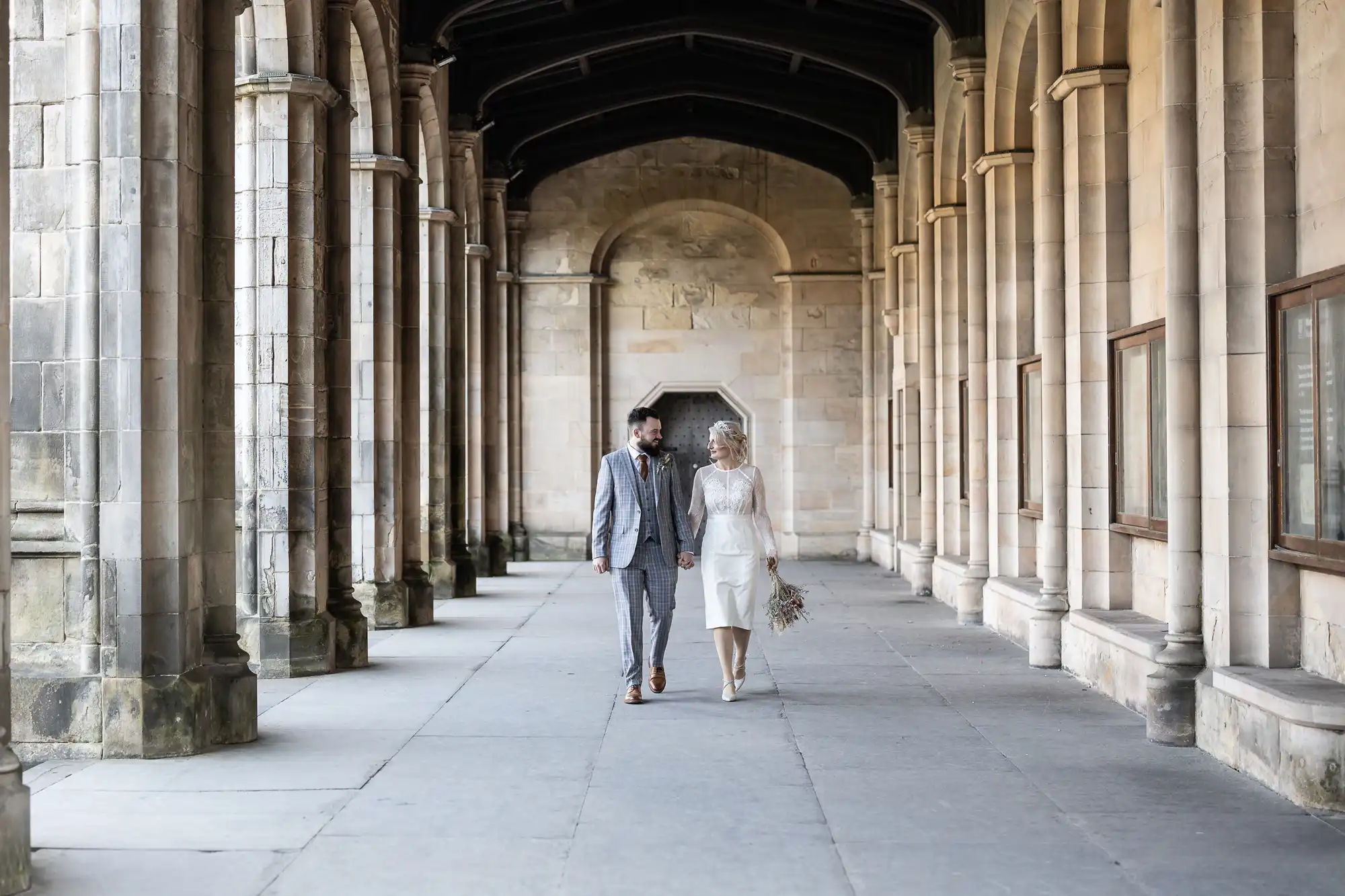 A couple walk hand-in-hand down a stone corridor with arched columns. The man wears a grey suit; the woman wears a knee-length white dress and holds a bouquet. Soft daylight filters in, creating a serene, romantic atmosphere in the historic setting.