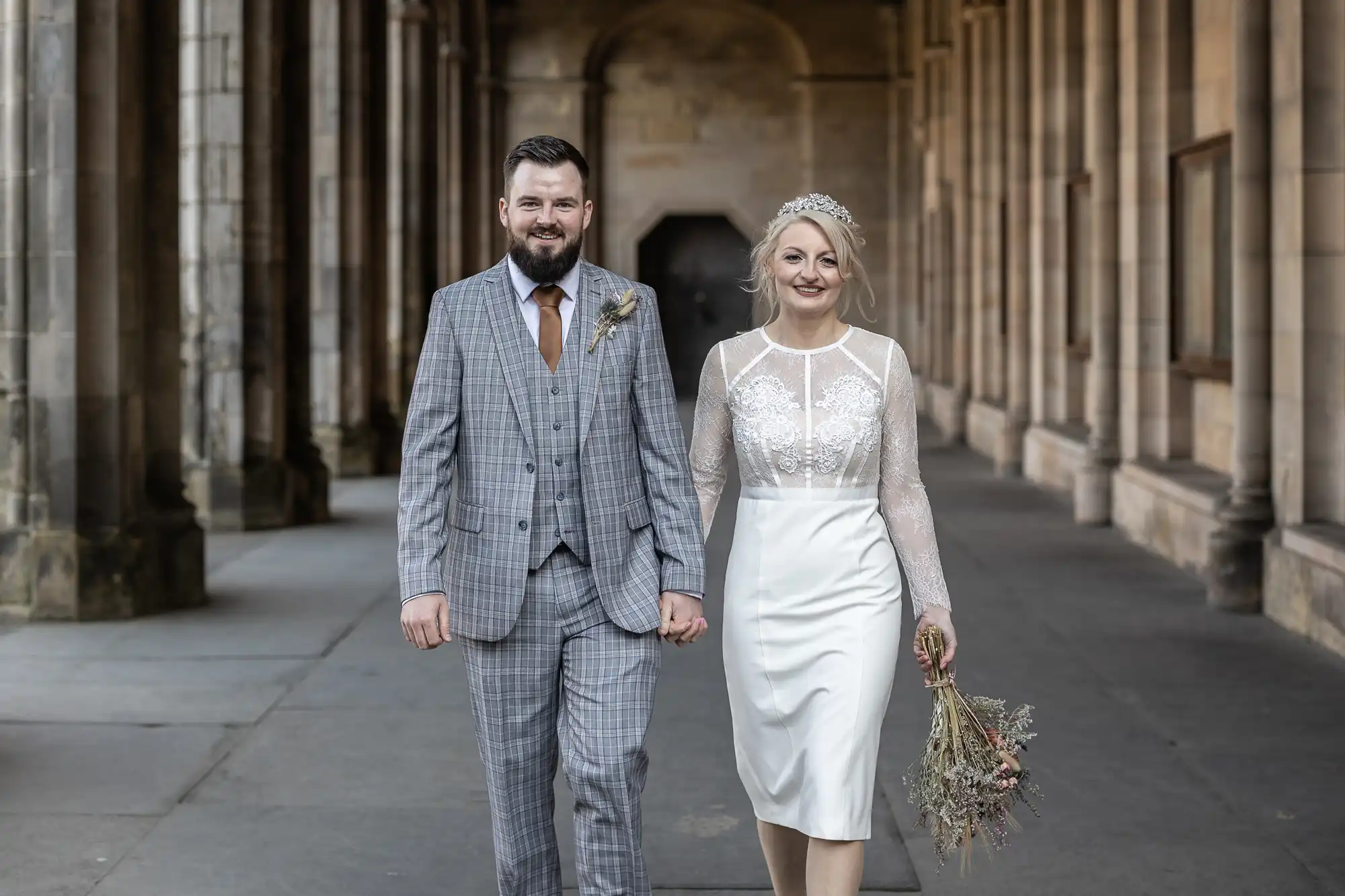 A smiling couple walk hand-in-hand down a stone corridor. The groom wears a grey checked suit and brown tie; the bride wears a white lace dress, a tiara, and holds a small bouquet of dried flowers. Arched columns and soft lighting frame the scene.