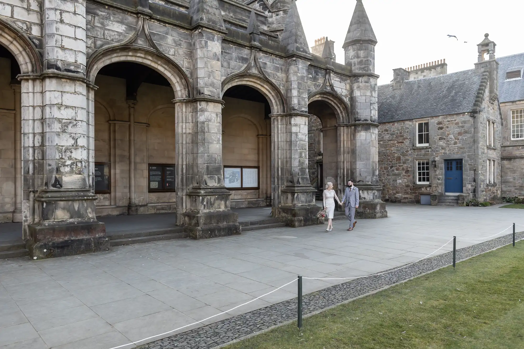 Two people walk side by side past a historic stone building with arched doorways and pointed towers. Noticeboards are visible under the arches, and another stone building with a blue door stands to the right on a paved courtyard.