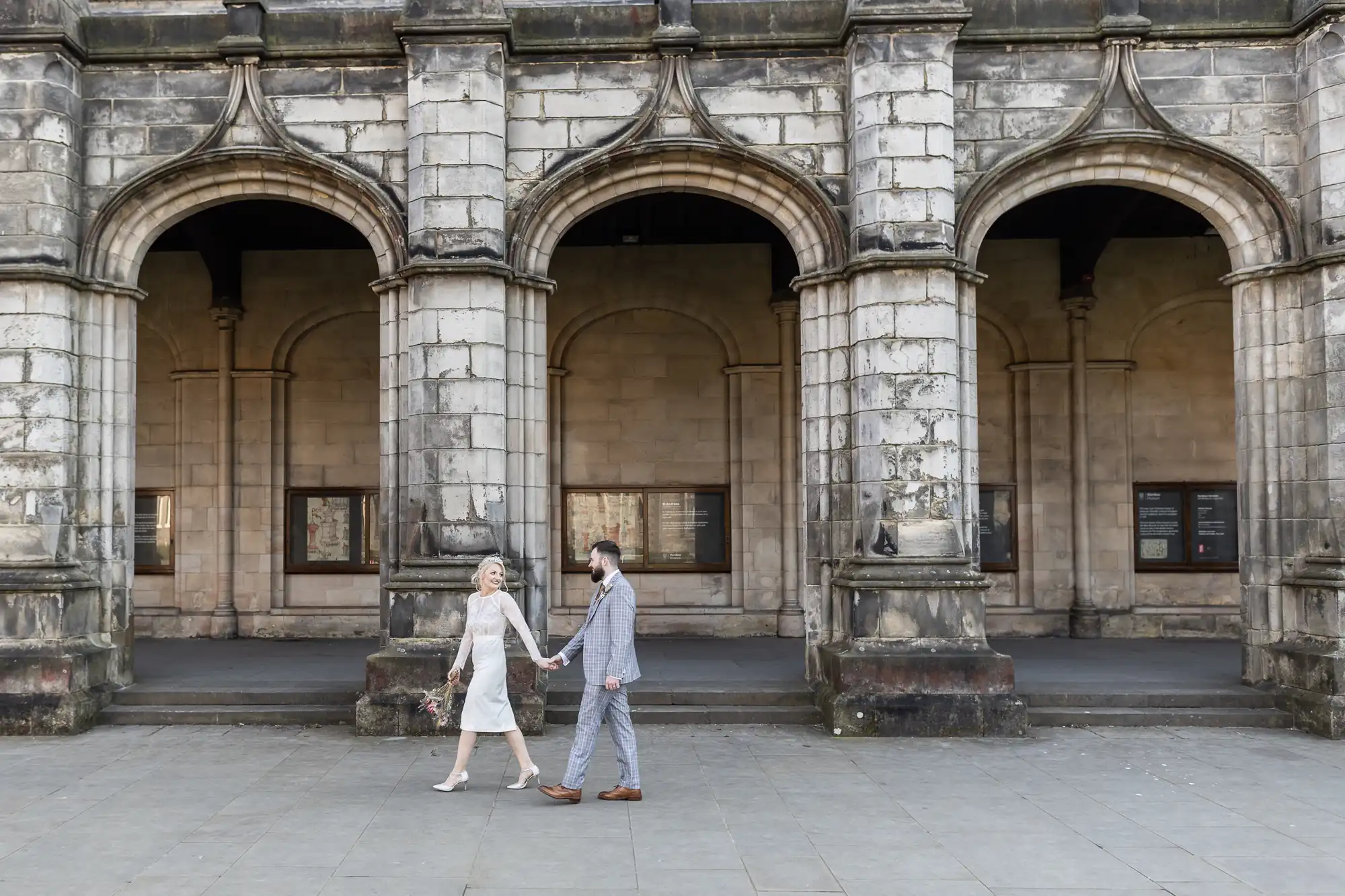 A couple dressed in wedding attire hold hands while walking past a historic stone building with three large arched doorways. The bride wears a white dress and holds a bouquet; the groom wears a checked suit. The scene is bright and elegant.