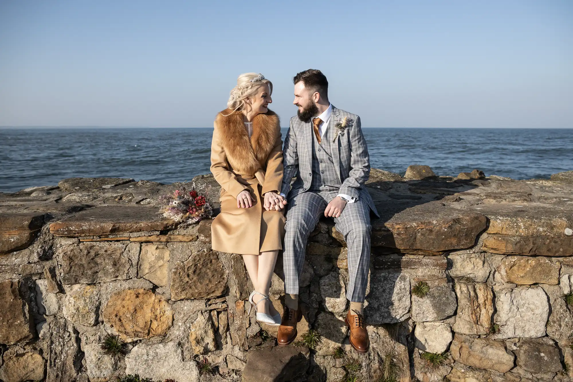 A couple dressed in formal attire sit on a stone wall by the sea, holding hands and smiling at each other. The woman wears a tan coat with a fur collar and silver heels; the man wears a grey checked suit with a brown tie. A bouquet rests beside them.
