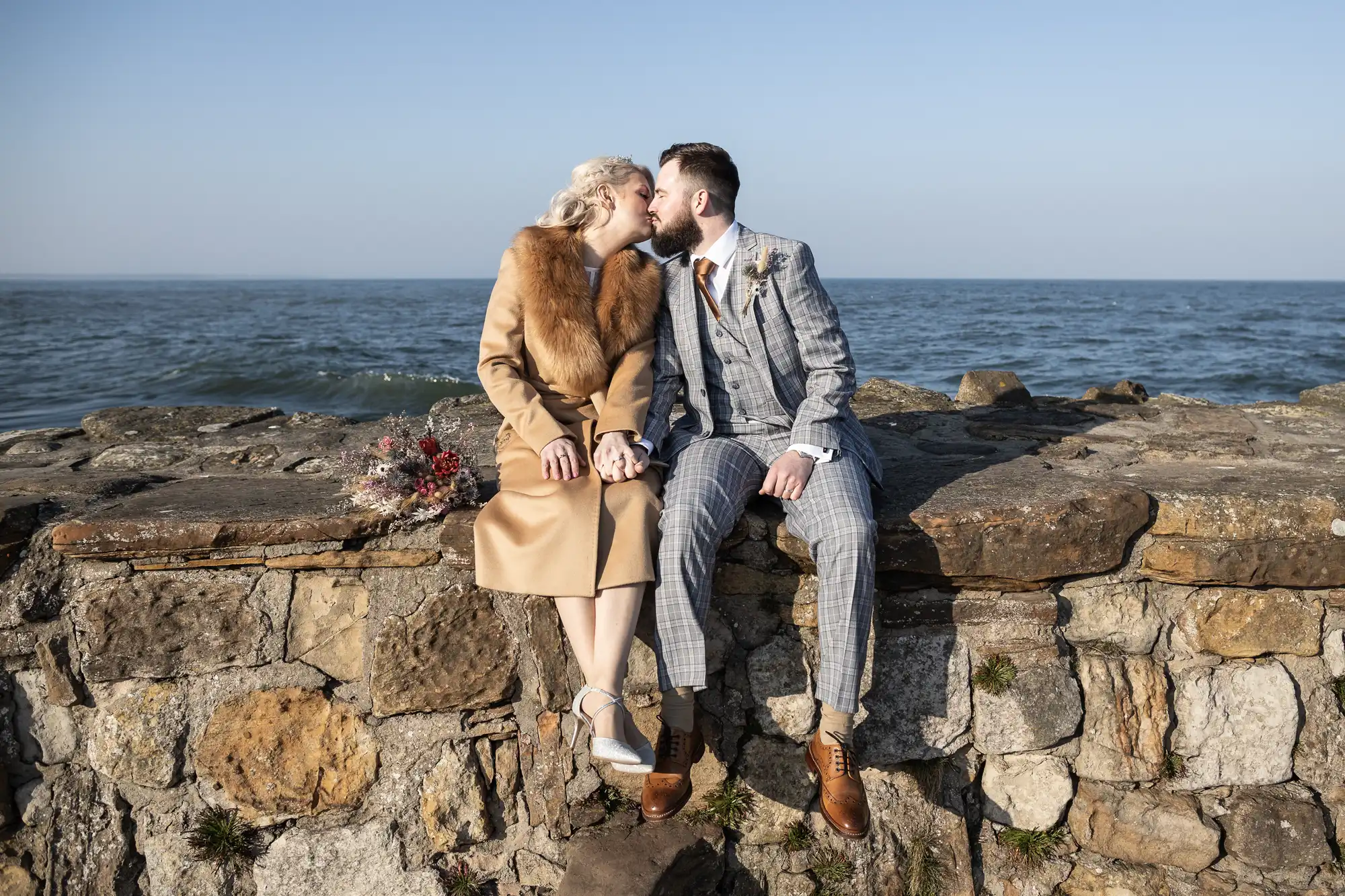 A couple sits on a stone wall by the sea, kissing. The woman wears a tan coat with a fur collar and silver heels, while the man wears a checked suit and brown shoes. A bouquet lies beside them. The sky is clear and the water is calm in the background.
