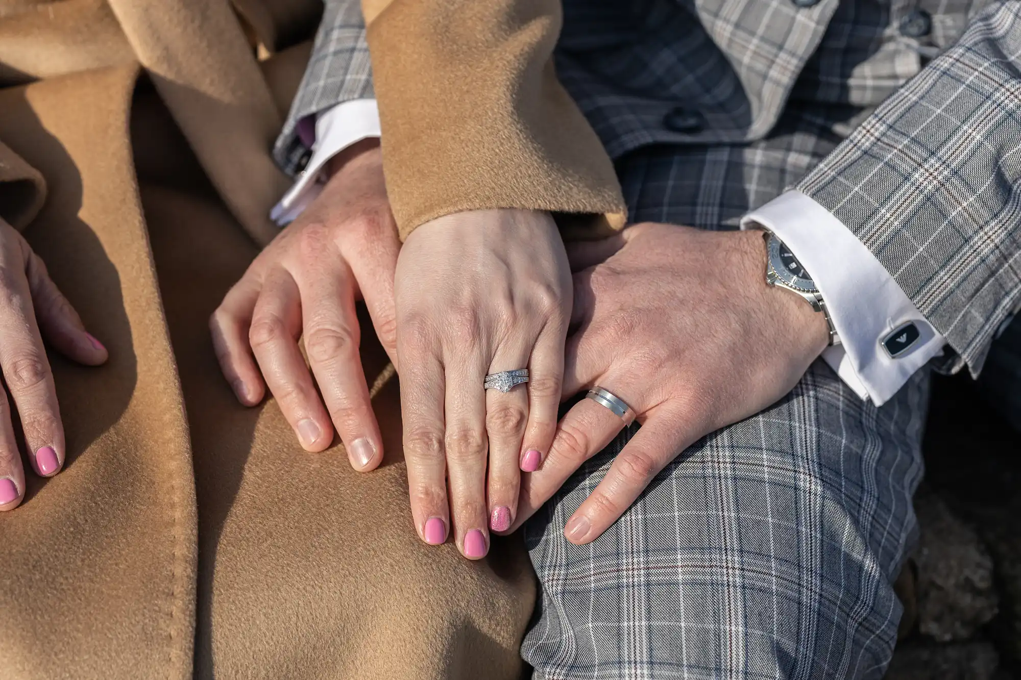 A close-up of two hands with silver rings resting on each other. Both people wear tan and grey checked coats. The person on the left has pink nail varnish and a diamond ring; the person on the right wears a watch and a plain silver band.
