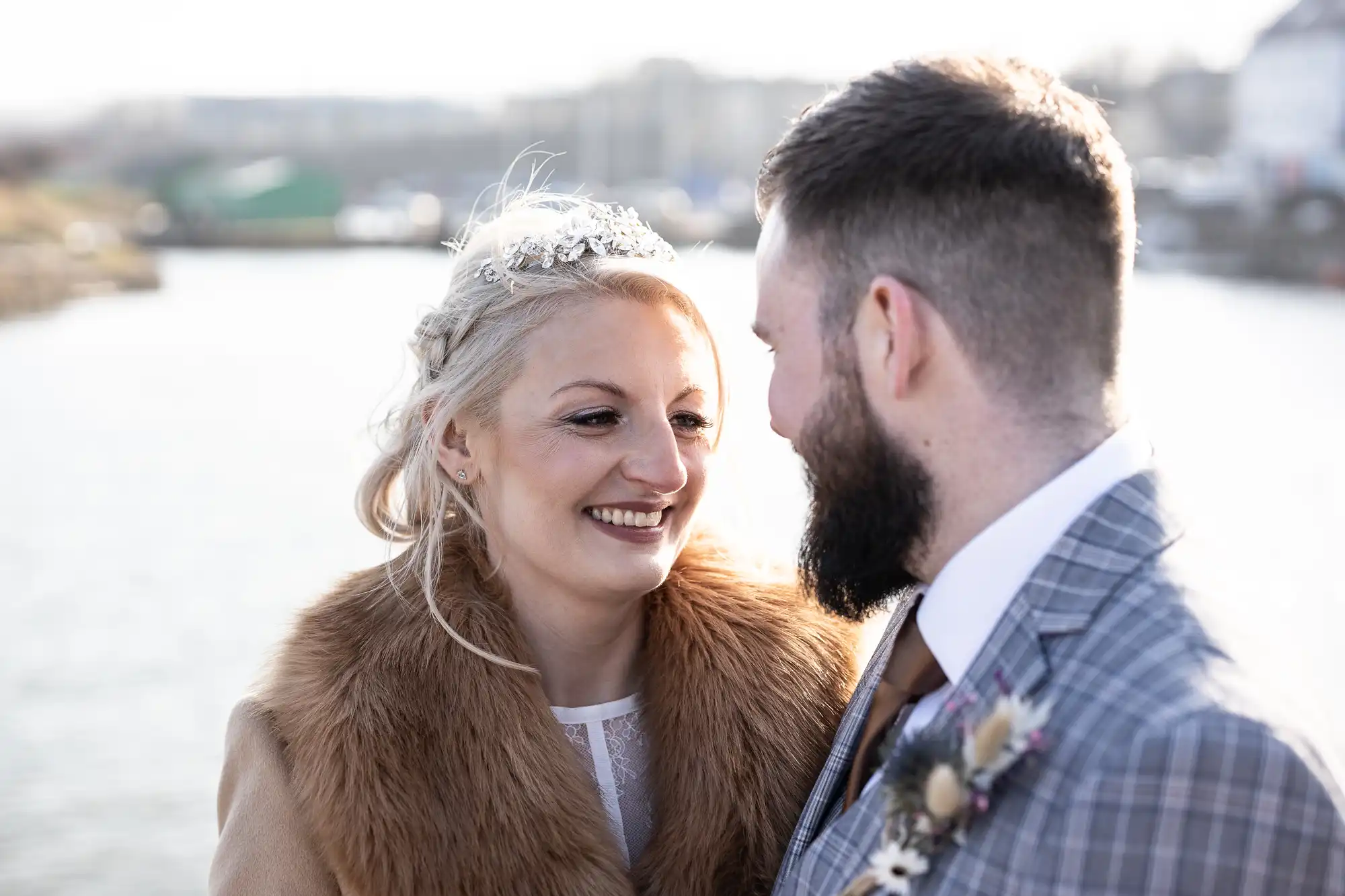 A smiling woman with blonde hair, a tiara, and a brown fur stole stands close to a bearded man in a grey checked suit. They face each other outdoors, with soft daylight and a blurred waterfront background.