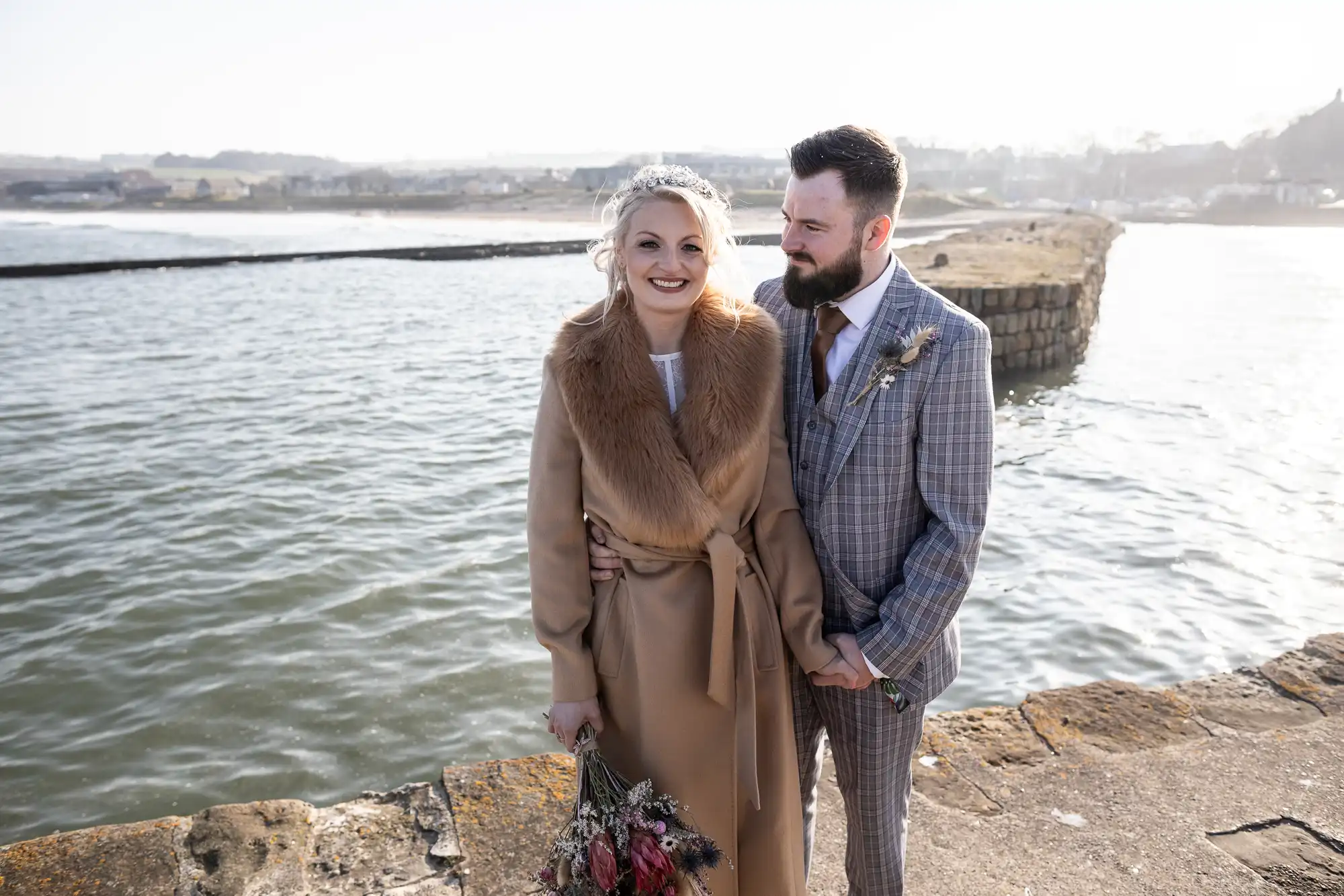 A smiling couple stand near a waterfront on a stone path. The woman wears a tan coat with a faux fur collar and holds a bouquet of dried flowers. The man, with a beard, wears a grey checked suit. The background features calm water and a hazy distant shoreline.