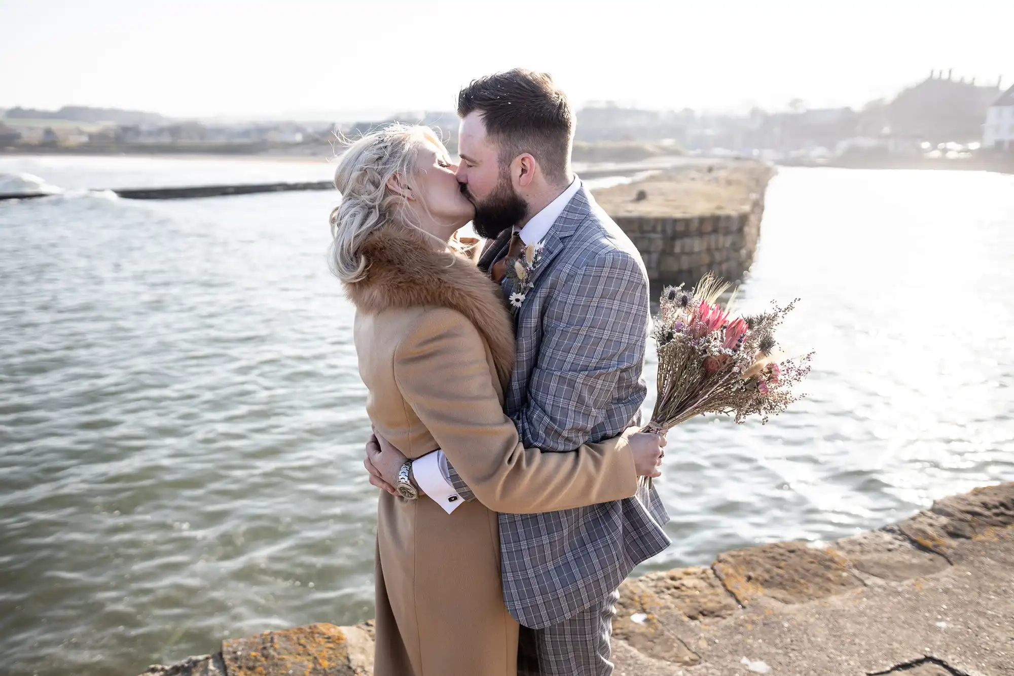 A couple stands on a stone path by the water, embracing and kissing. The woman wears a tan coat with a fur collar and holds a bouquet of wildflowers. The man wears a checked grey suit. The scene is bright and outdoors, with a blurred shore in the background.