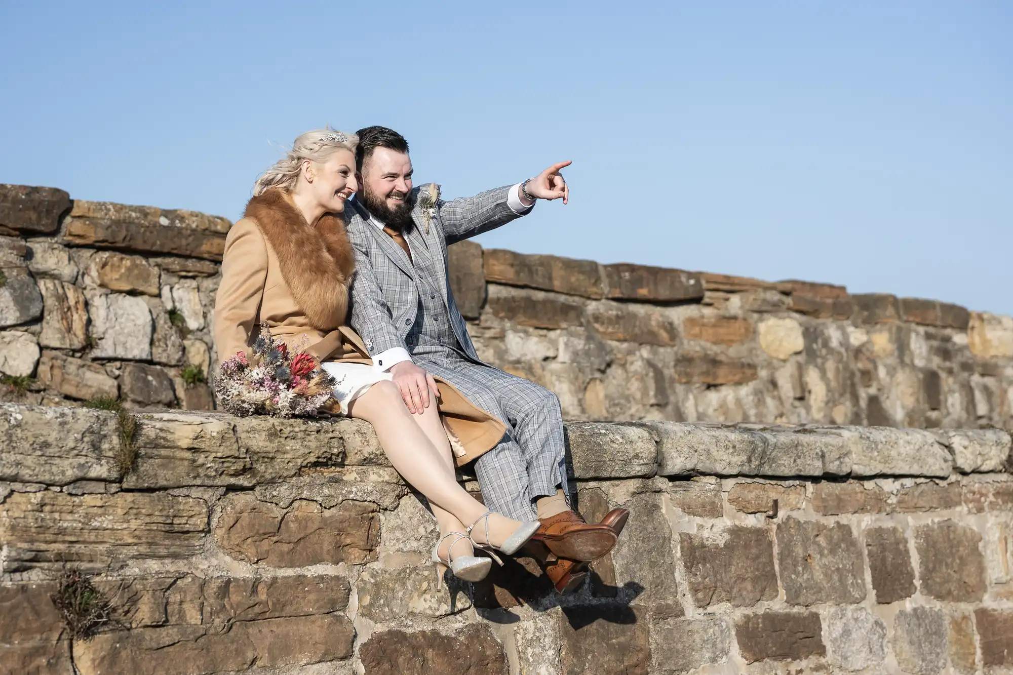 A smiling couple sits on a stone wall under a clear sky; the woman in a tan coat with a fur collar and holding a bouquet, the man in a grey checked suit points into the distance, both appearing happy and relaxed outdoors.