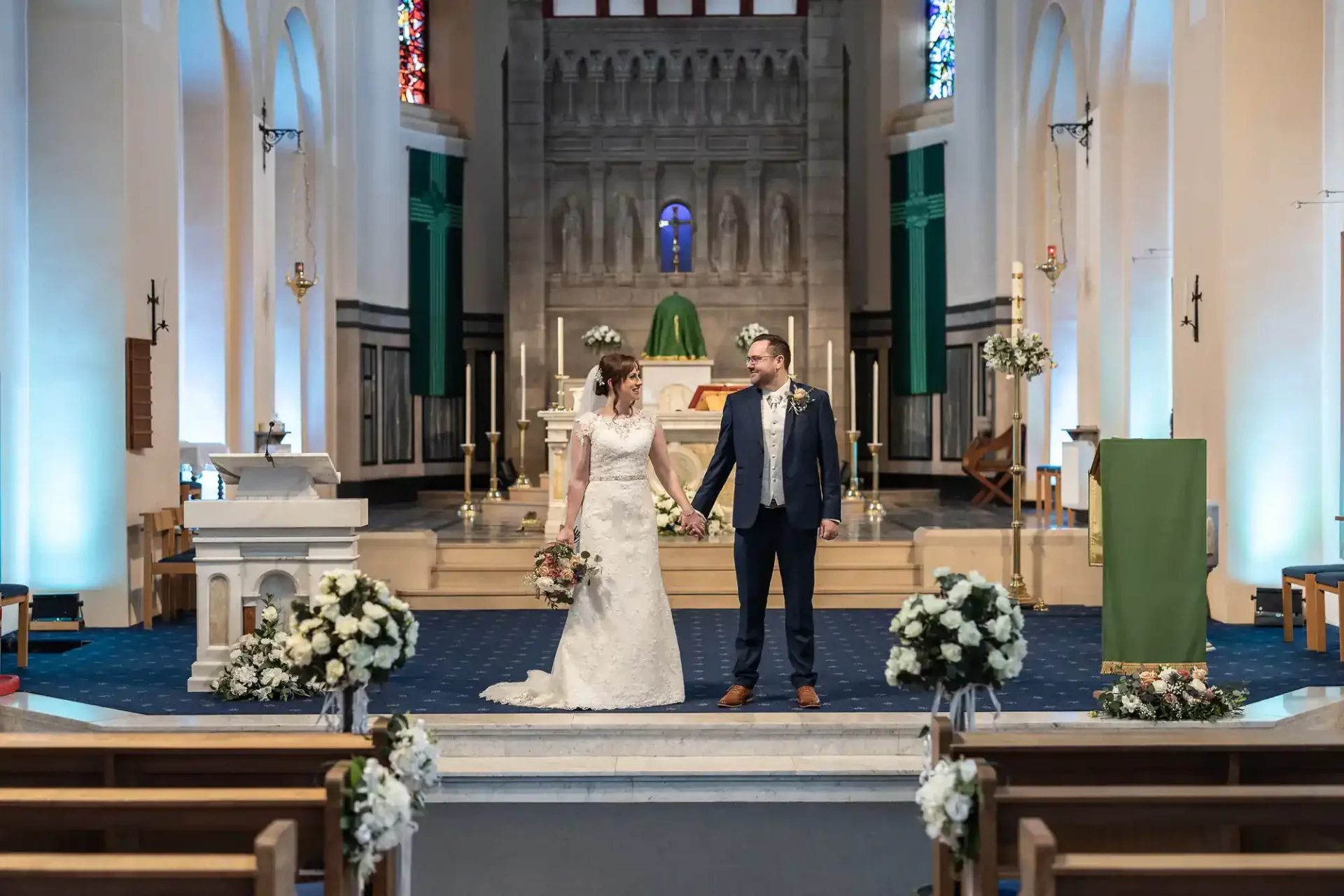 A bride in a white dress and a groom in a navy suit hold hands, standing at the altar of a decorated St Margaret’s Memorial Church in Dunfermline. White flower arrangements line the aisle, and soft blue lighting illuminates the arched interior and stained glass windows.