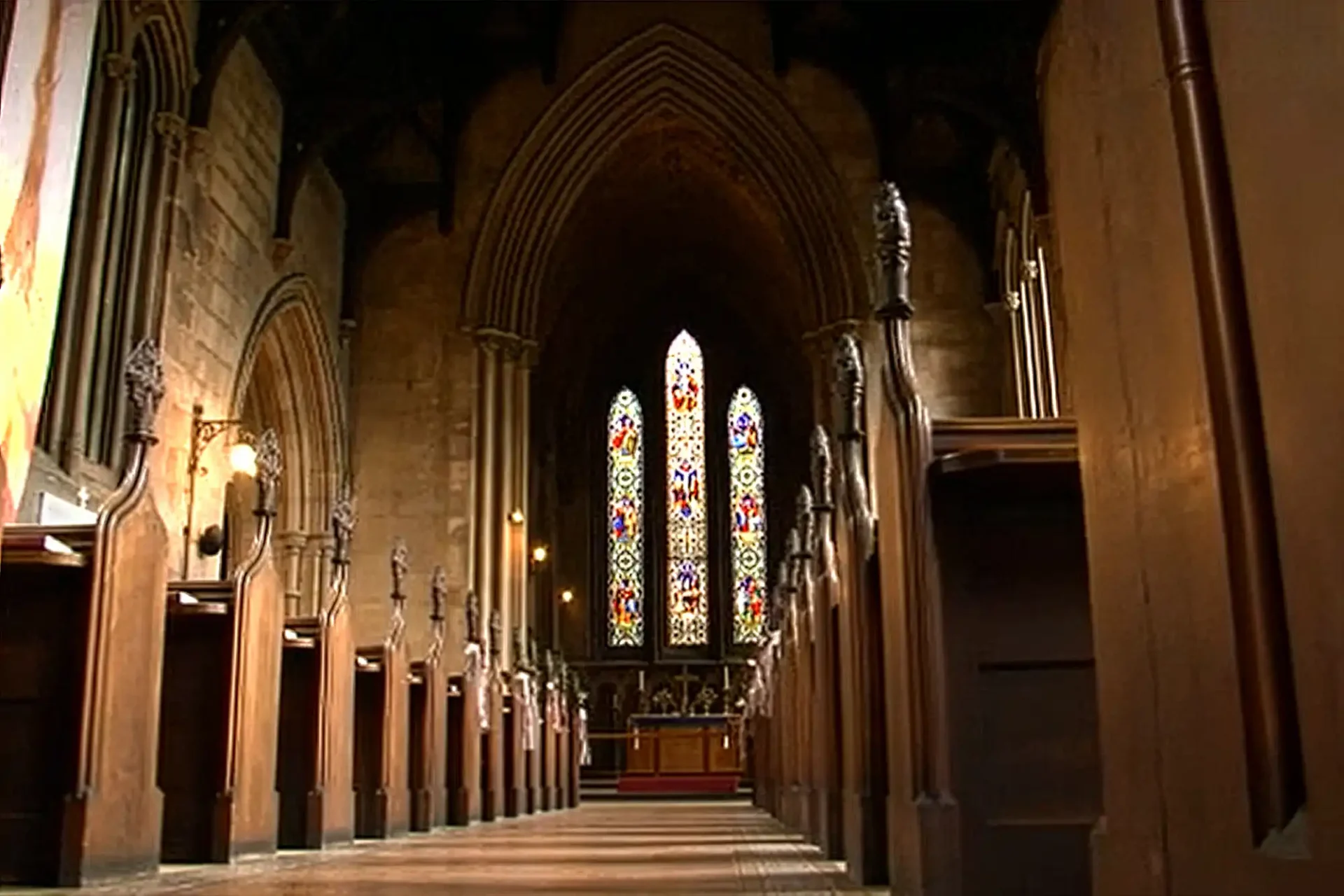 A view down the aisle of a gothic St Mary's Scottish Episcopal Church, Dalkeith interior with high vaulted ceilings, wooden pews on each side, and three tall, ornate stained glass windows behind an altar at the far end, letting in colourful light. The scene is empty and peaceful.