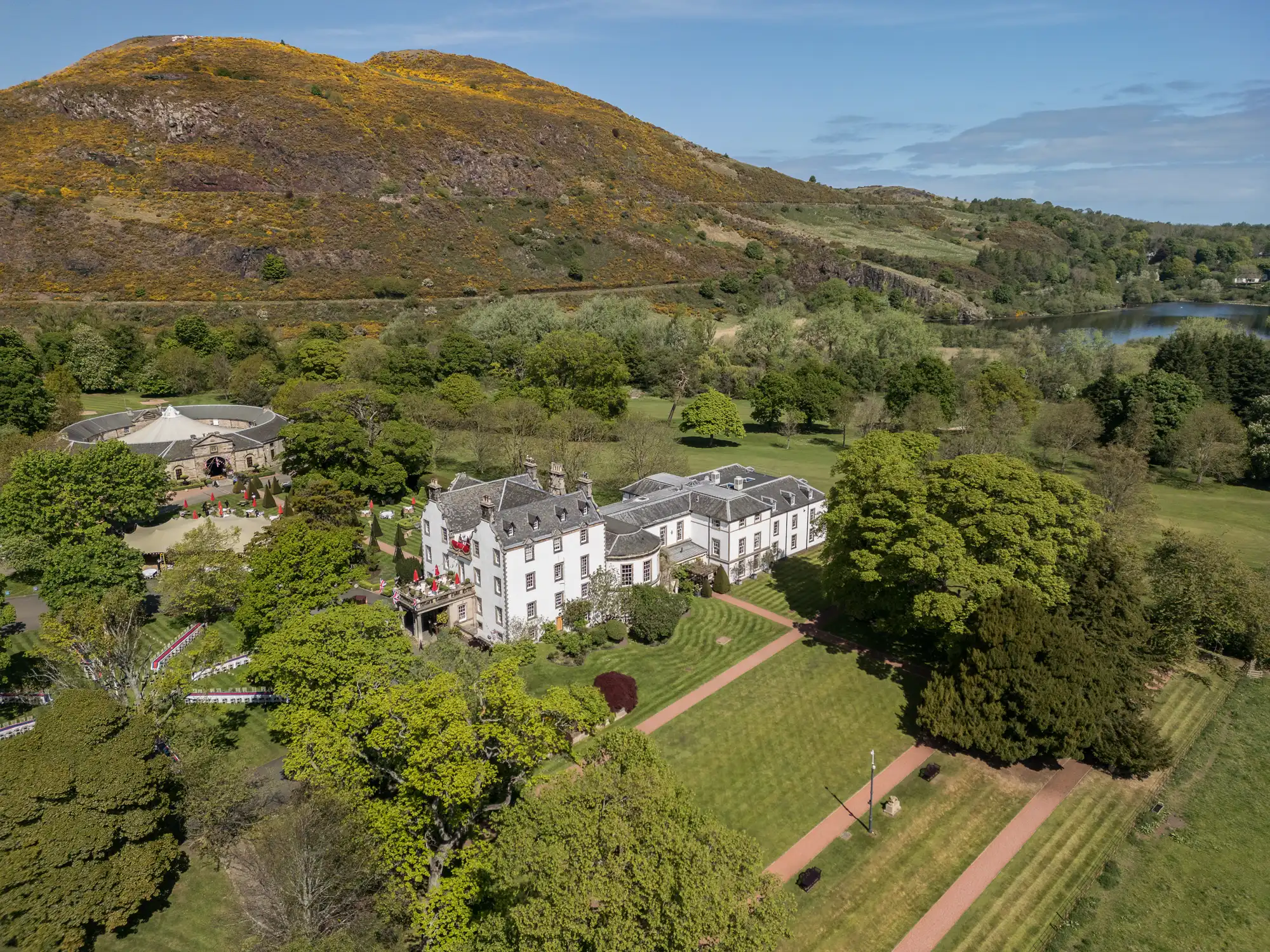 A grand white mansion surrounded by lush green lawns and large trees sits at the base of a hill covered in shrubs and yellow wildflowers, with a lake and more greenery visible in the distance under a blue sky.
