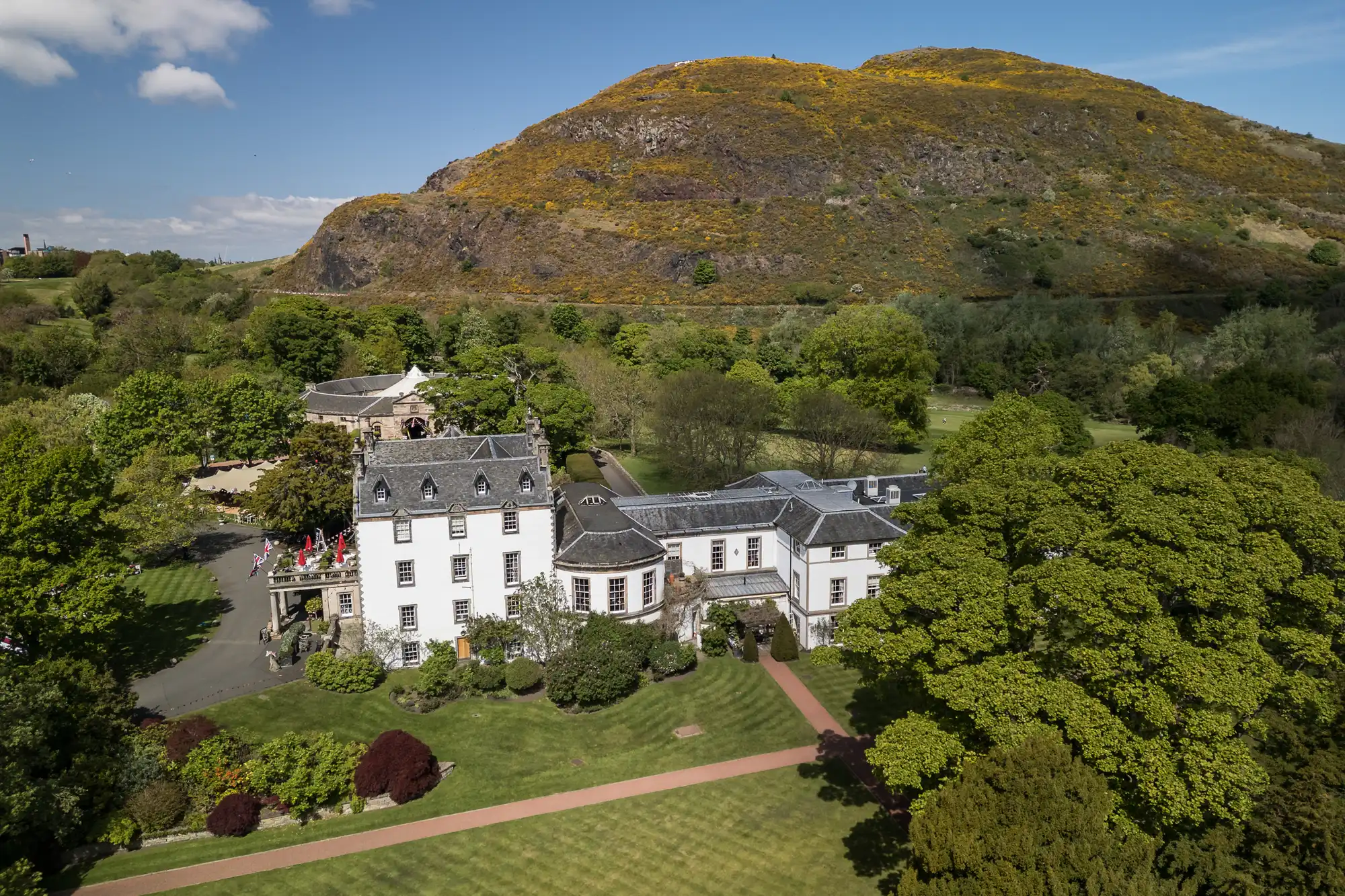Aerial view of a large white historic building surrounded by gardens and green trees. In the background, a steep, grassy hill rises under a blue sky with scattered clouds. Pathways and manicured lawns are visible around the building.