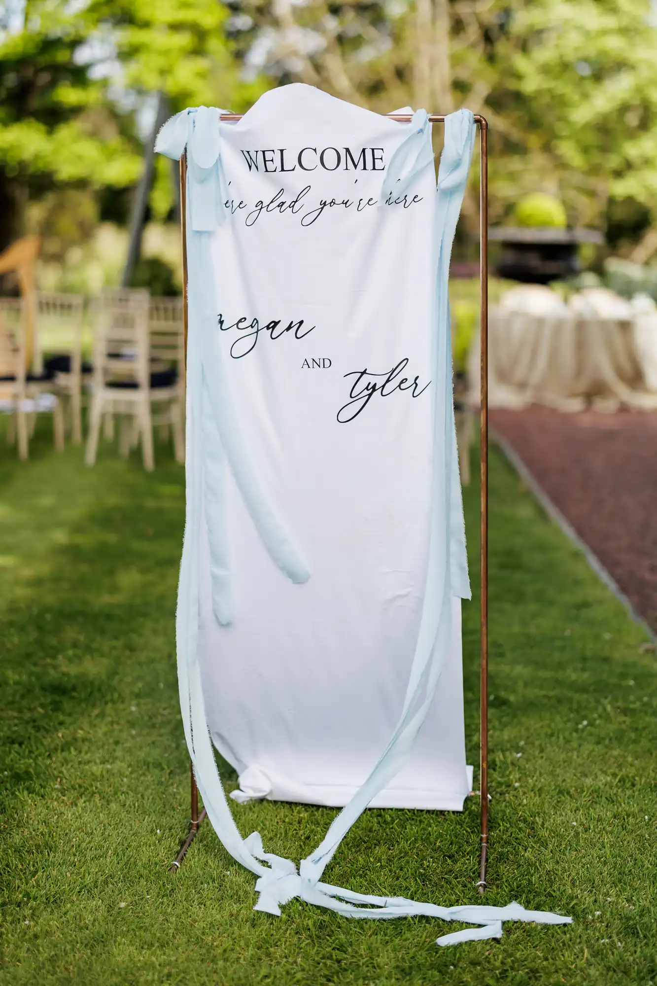 A white fabric sign with flowing blue ribbons hangs outdoors, welcoming guests with black script text reading "Welcome, we’re glad you’re here, Regan and Tyler." The sign stands on grass with blurred chairs and greenery in the background.