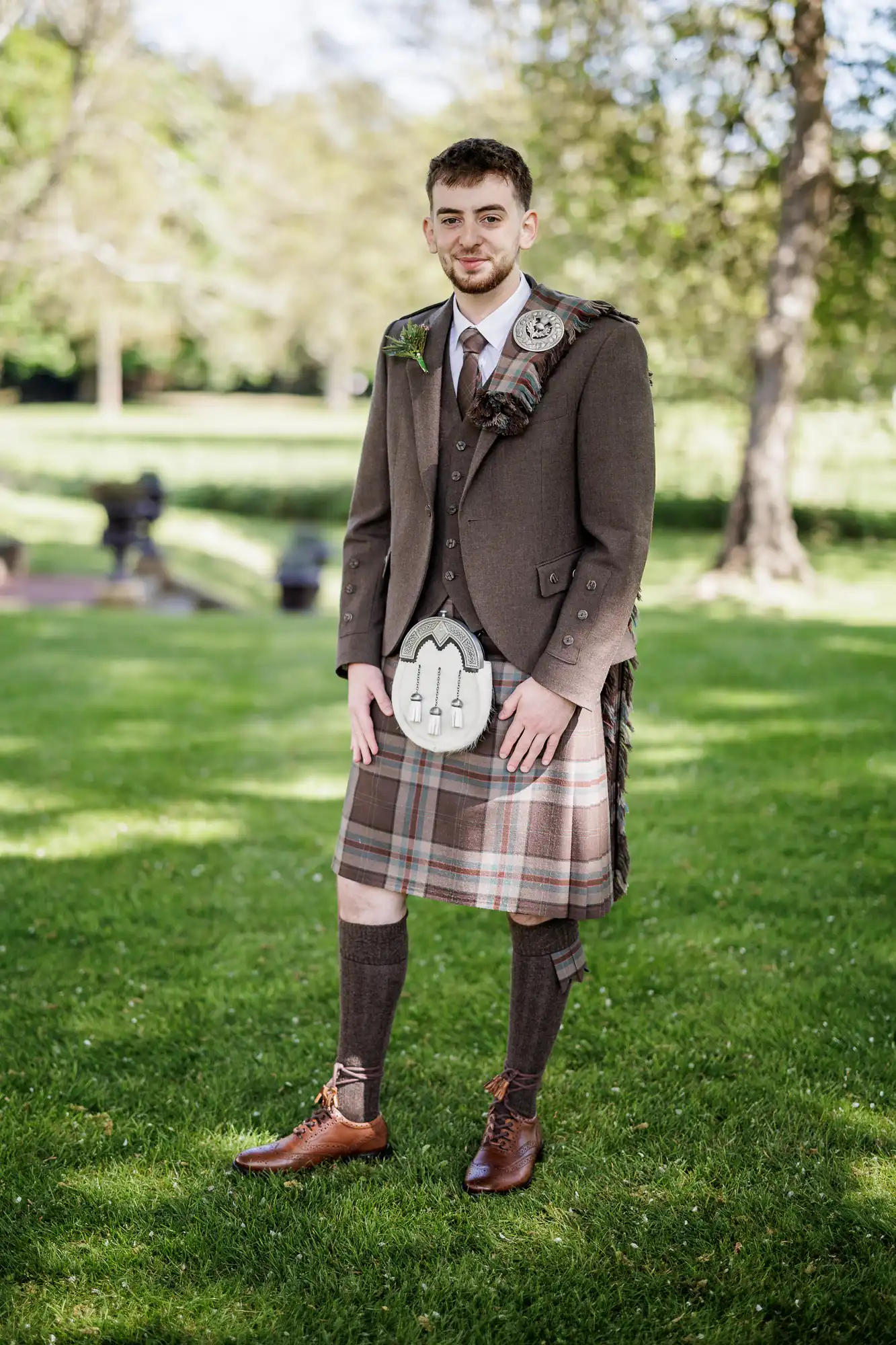 A young man stands on grass wearing a traditional Scottish outfit: a brown jacket, tartan kilt and sash, knee-high socks, brown shoes, a sporran, and a buttonhole. The background is bright and leafy with sunlight filtering through trees.