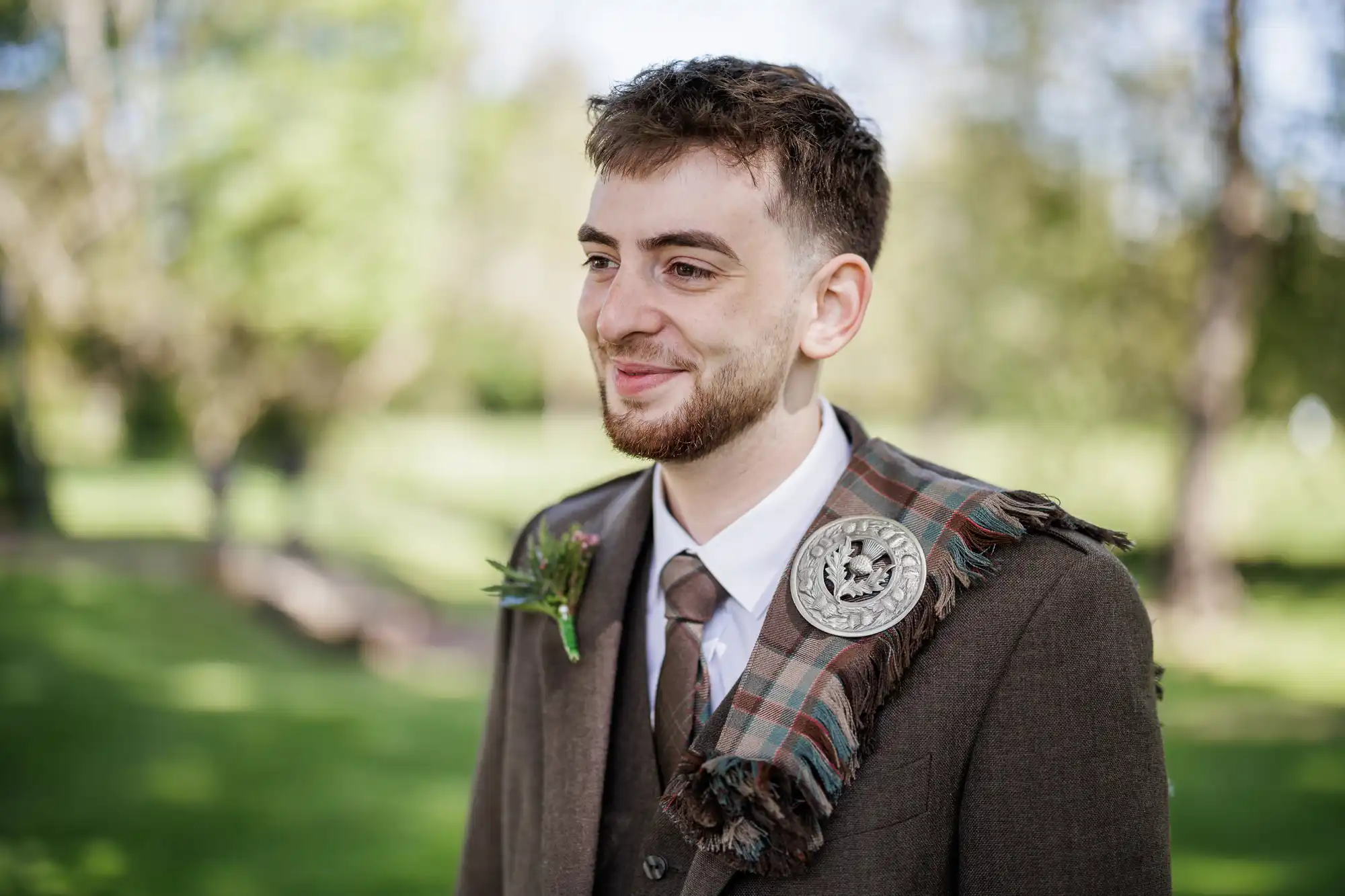 A young man with short brown hair and a beard smiles outdoors, wearing a brown suit with a tartan sash and a decorative silver brooch. He has a buttonhole on his lapel. The background is green and blurred, suggesting a natural, sunny setting.