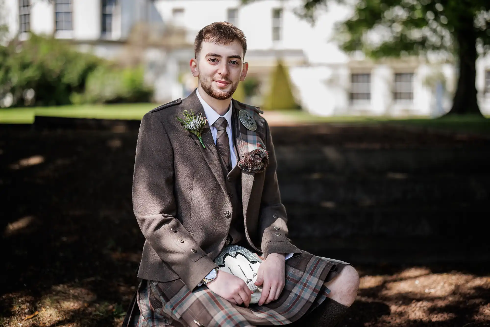 A young man sits outdoors on a stone ledge, wearing a traditional brown Scottish kilt outfit with a sporran and a buttonhole on his jacket. He smiles gently, with trees and a white building blurred in the sunlit background.