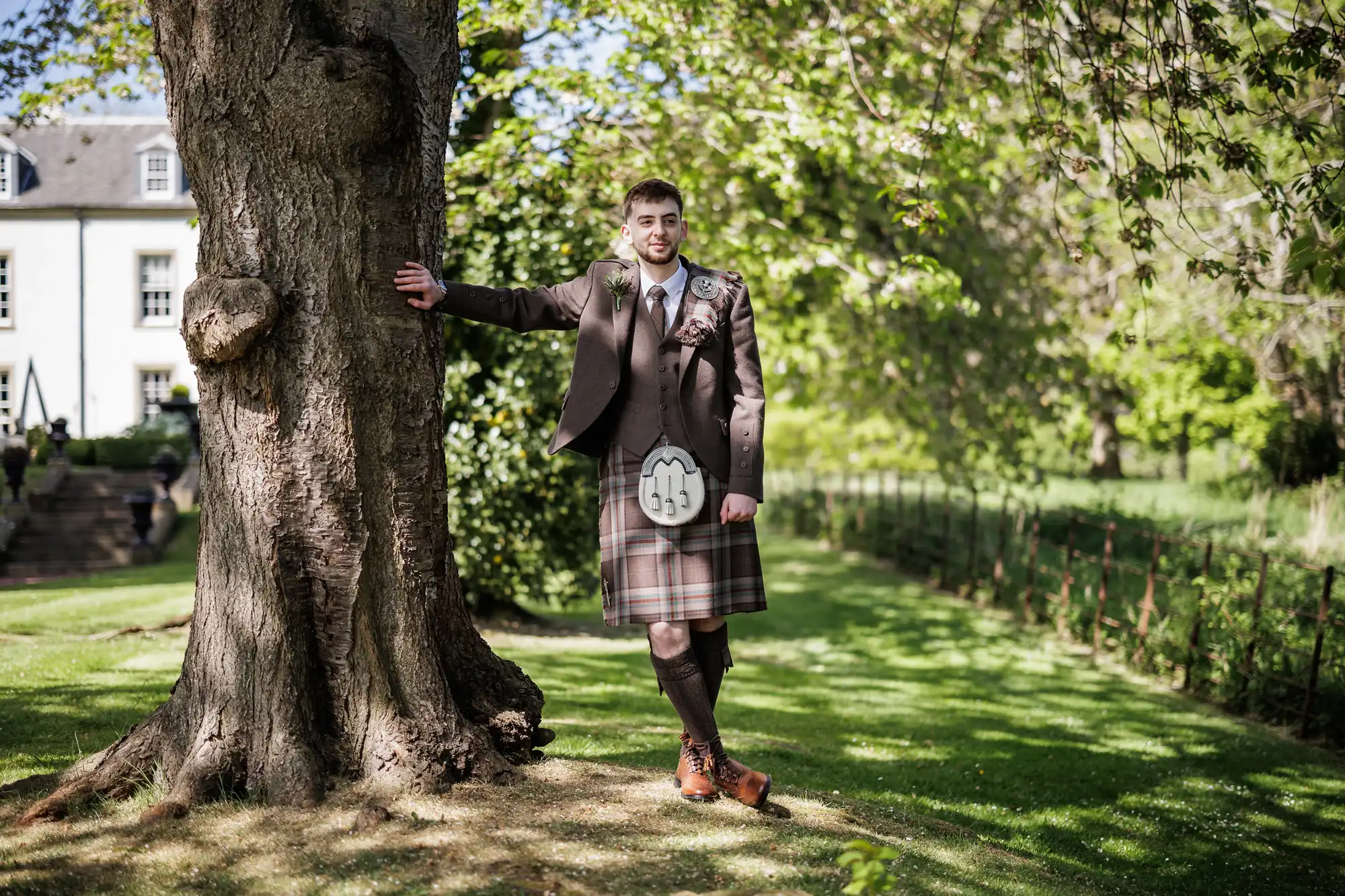 A man in a brown checked kilt, brown jacket, and tie stands outdoors, leaning on a large tree trunk. He is smiling, surrounded by lush green grass and trees, with a white building visible in the background under bright daylight.