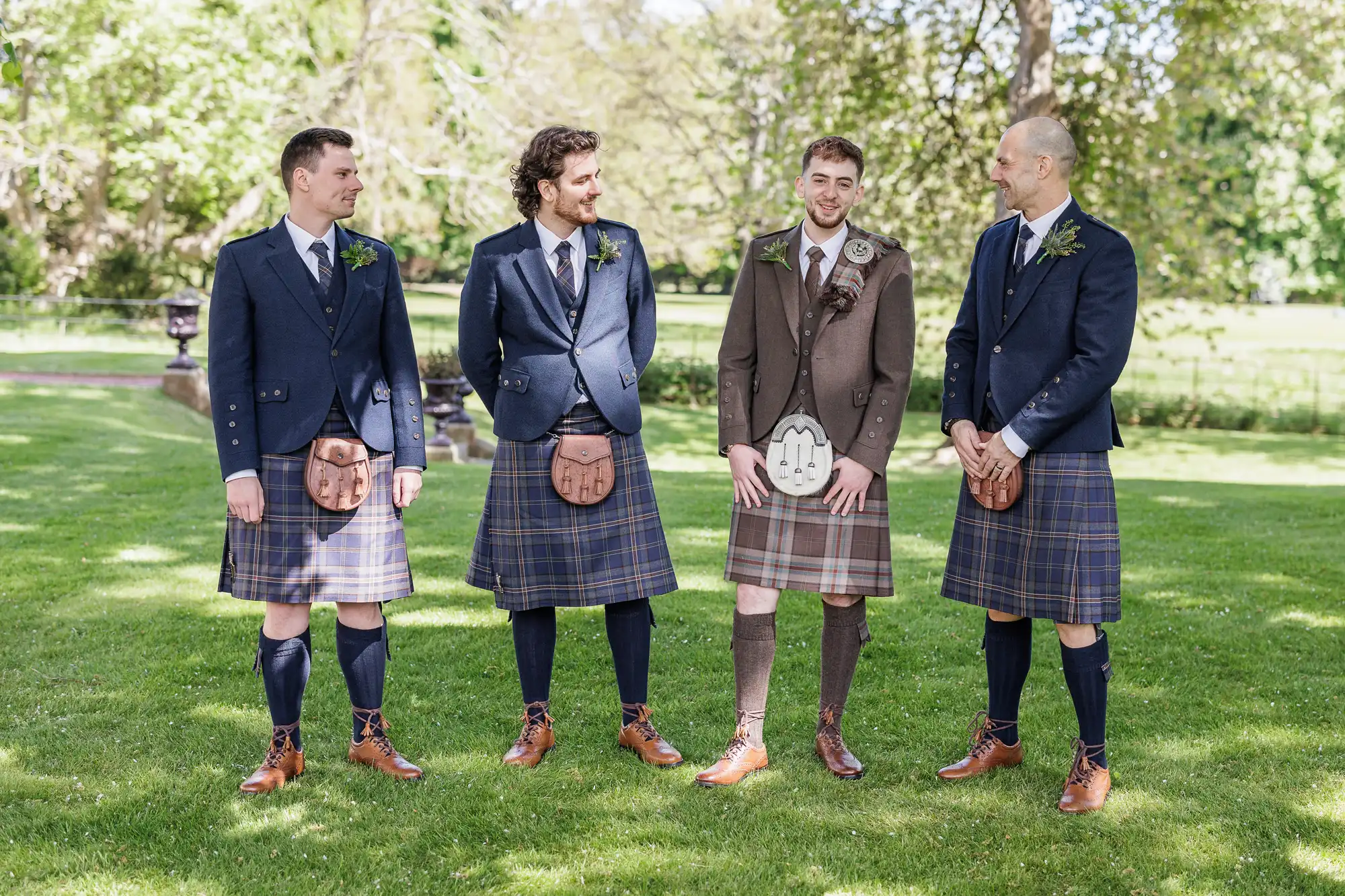 Four men stand on grass, smiling and talking. Three wear blue jackets, plaid kilts, sporrans, knee socks, and brown shoes. One, likely the groom, wears a brown jacket and kilt with a white sporran. Trees and sunlight fill the background.
