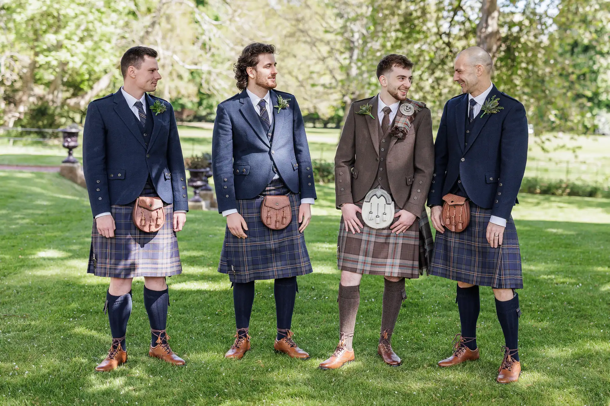 Four men stand on grass wearing kilts, dress shoes, knee-high socks, shirts, and jackets. Three wear navy jackets and blue tartan kilts, whilst one wears a brown jacket and brown tartan kilt. They all smile at each other with trees and sunlight in the background.