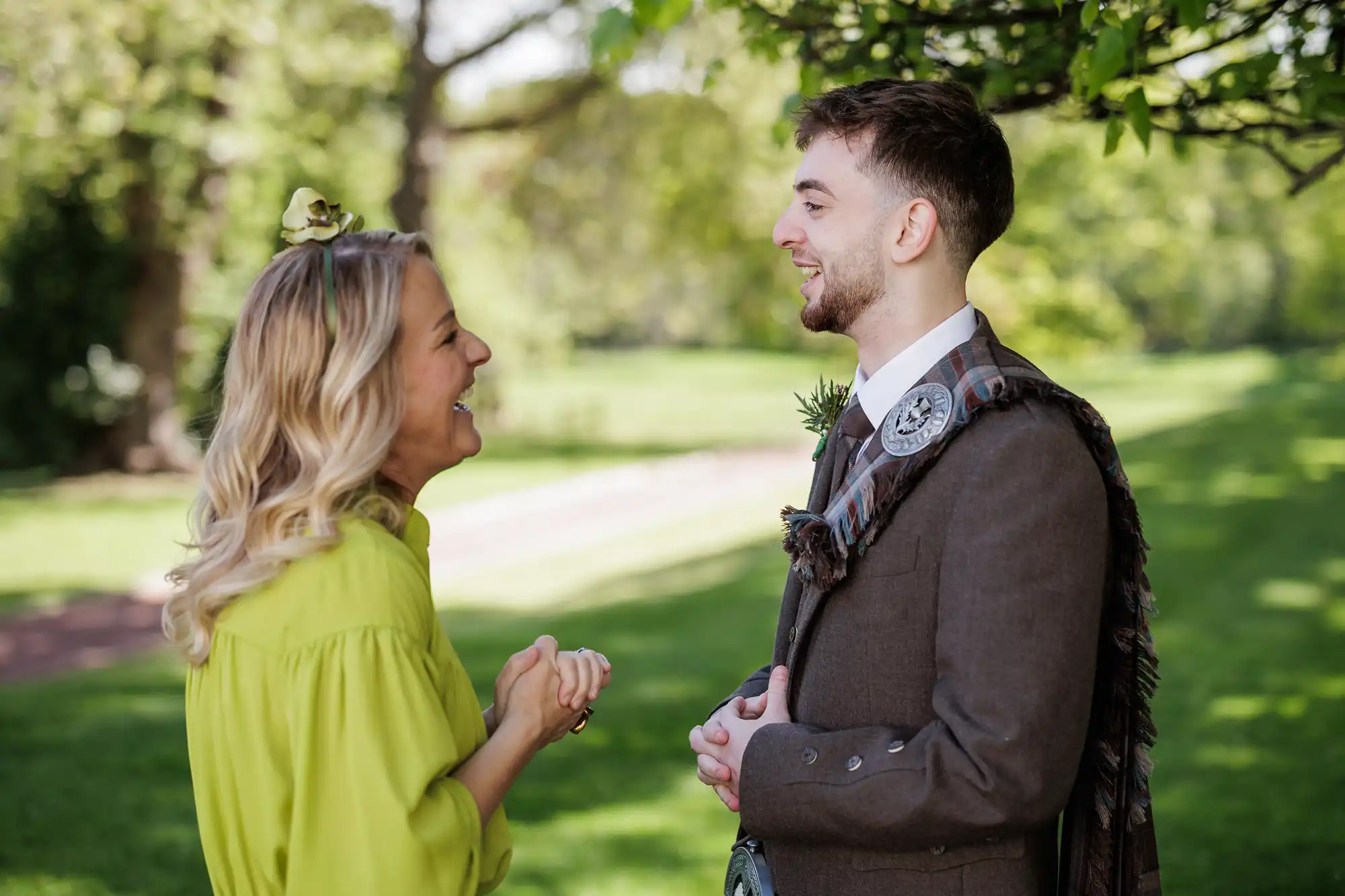 A woman in a bright green dress laughs as she talks to a man in a brown suit and tartan sash, standing outdoors under leafy trees on a sunny day. Both are smiling and appear to be enjoying a pleasant conversation.